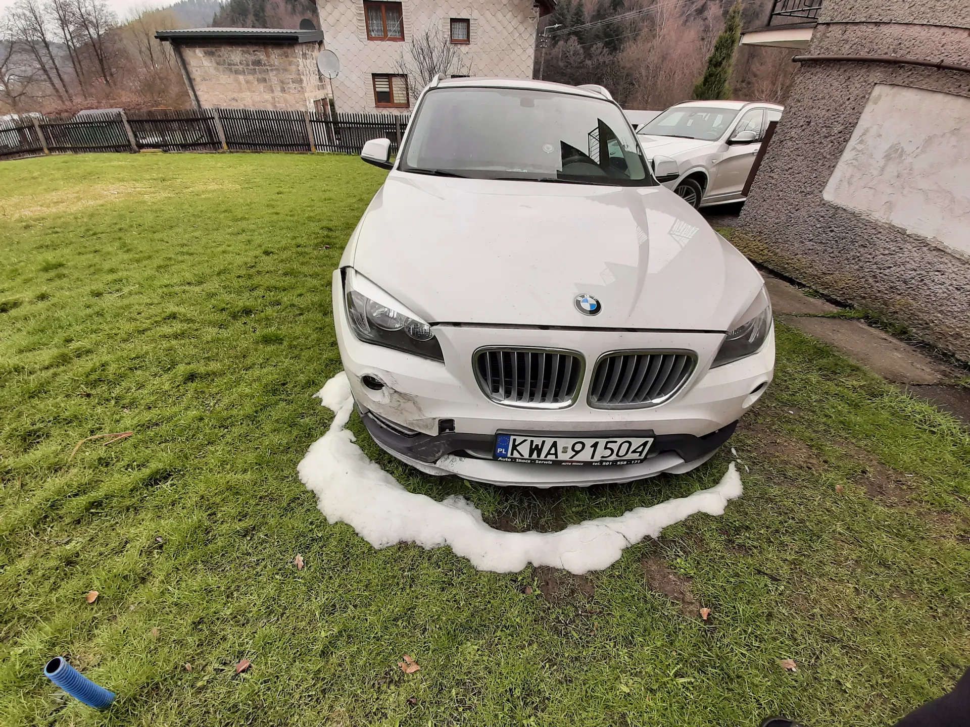Biały samochód marki BMW z tablicą rejestracyjną PL KWA-91504, zaparkowany na trawie. Przedni zderzak po lewej stronie uszkodzony. Wokół przedniej części pojazdu widoczny śnieg w kształcie półkola. Na tle drugi biały samochód, dom z kamiennej ściany, ogrodzenie, drzewa oraz niebieska rura na trawie. Trawa zielona, na niej rozrzucone suche liście.