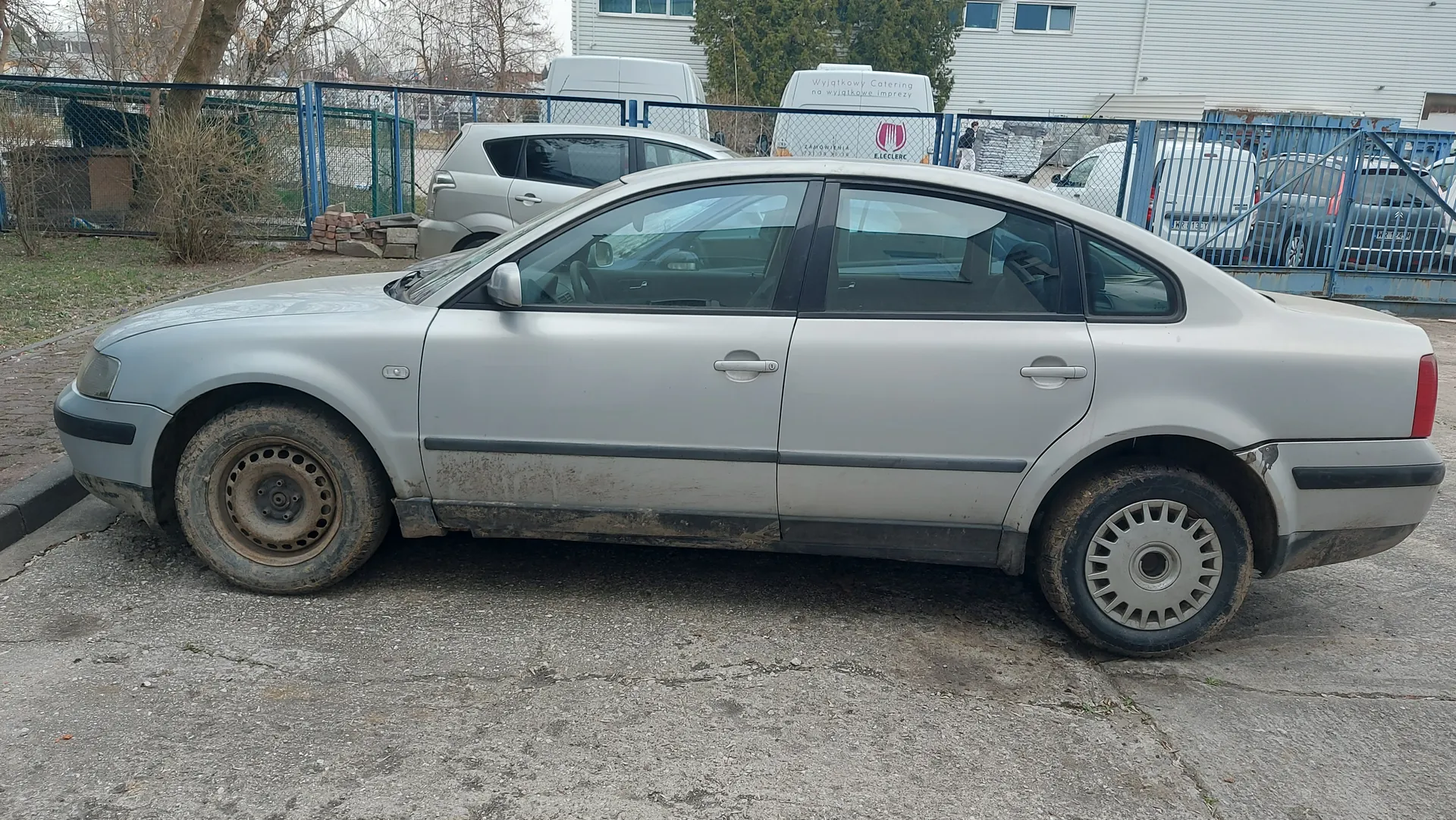 Srebrny sedan zaparkowany na betonowej powierzchni, z widocznymi śladami brudu na karoserii i oponach. W tle biały van z napisem "Wyjątkowy Catering na wyjątkowe imprezy" oraz logo "E. Leclerc", niebieskie metalowe ogrodzenie, budynek z oknami, suche krzewy i stos cegieł. Inne pojazdy, w tym srebrny samochód osobowy i białe furgony, znajdują się za ogrodzeniem. Asfaltowa powierzchnia parkingu z widocznymi pęknięciami.