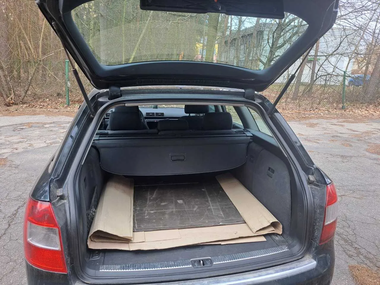 Czarny samochód typu kombi z otwartym bagażnikiem. Wewnątrz bagażnika leżą kawałki kartonu. Tło przedstawia parking z suchymi liśćmi, drzewami bez liści i budynkiem w oddali. Samochód stoi na asfaltowej nawierzchni.