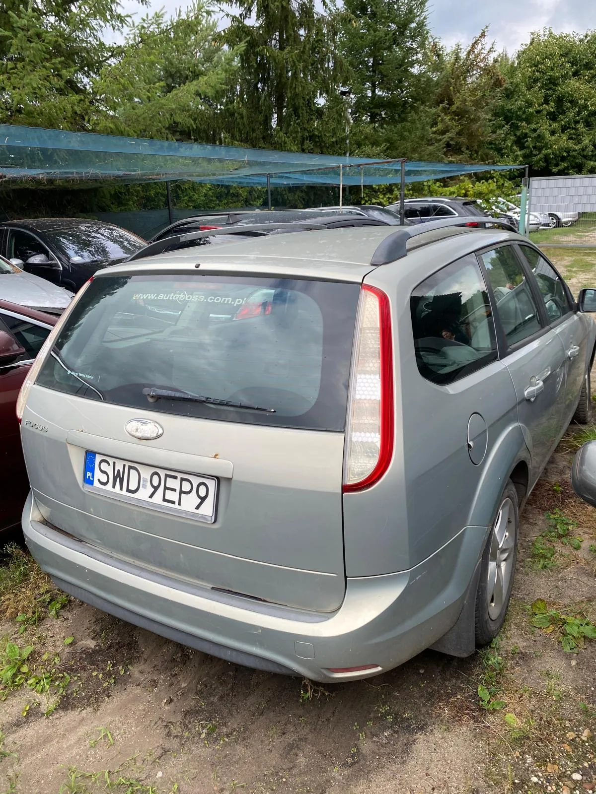 Tylny widok srebrnego samochodu typu kombi zaparkowanego na piaszczystym podłożu. Na tylnej szybie znajduje się naklejka z adresem strony internetowej www.autoboss.com.pl. Samochód posiada polską tablicę rejestracyjną o numerze SWD 9EP9. W tle widoczne są inne zaparkowane pojazdy, zielone drzewa oraz niebieska konstrukcja zadaszenia.