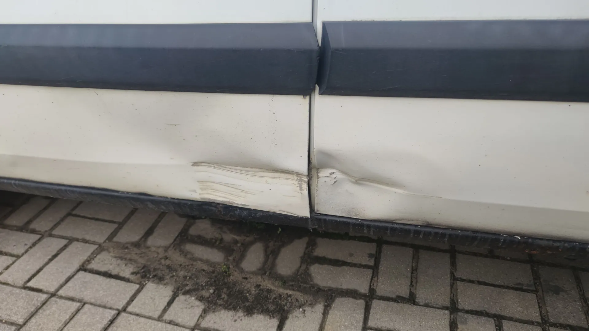 Zbliżenie na dolną część białego nadwozia samochodu. Widoczne są wgniecenia i zarysowania lakieru na drzwiach oraz progu, tuż nad czarną listwą ochronną. Samochód stoi na kostce brukowej.