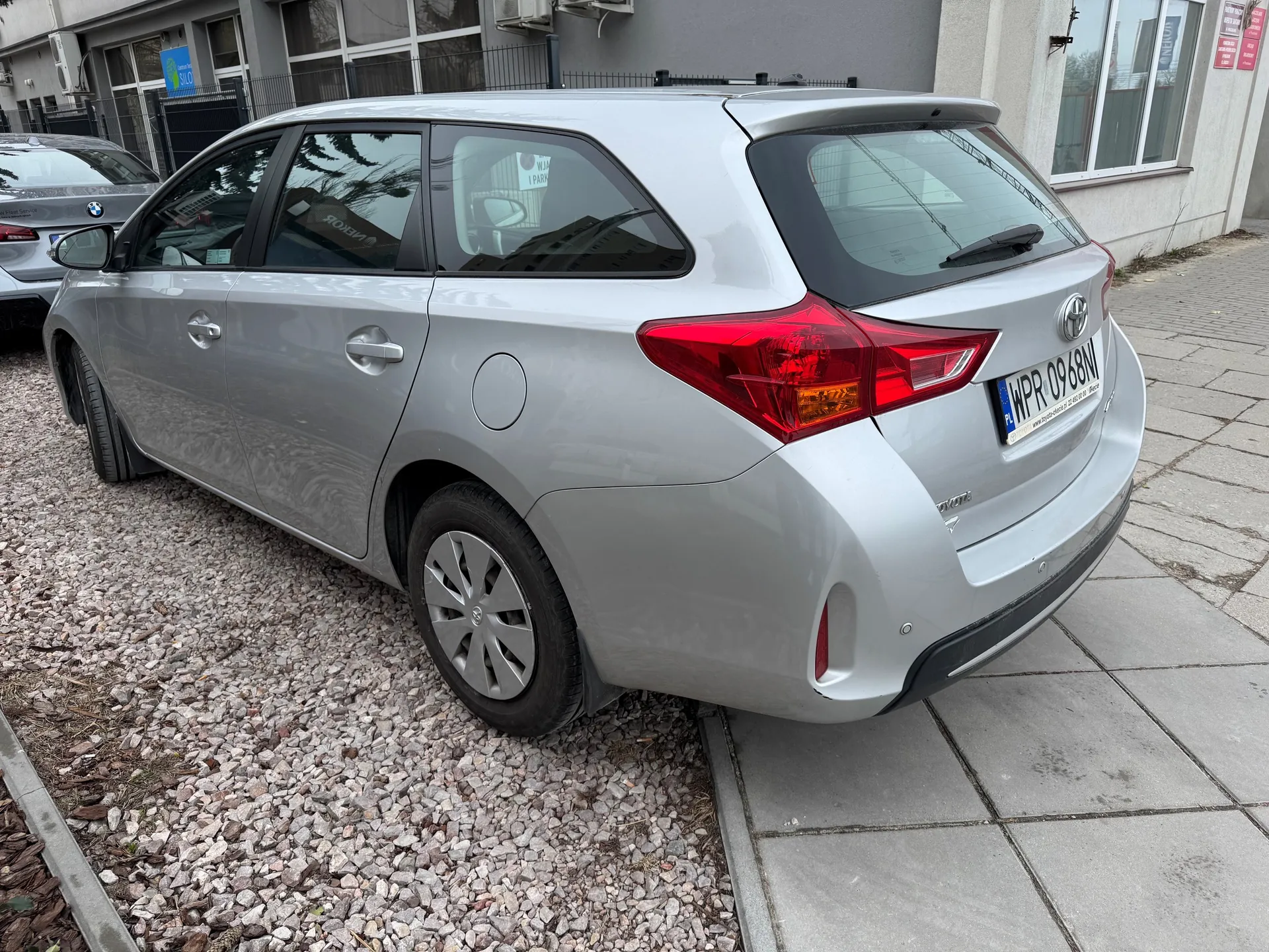 Srebrny samochód osobowy typu kombi marki Toyota zaparkowany na żwirowym obszarze obok chodnika z betonowych płyt. Widoczny tył pojazdu z czerwonymi światłami, tablicą rejestracyjną WPR 0968M oraz logo marki. W tle fragment budynku z oknami, ogrodzeniem i częściowo widocznym srebrnym samochodem marki BMW. Na szybie tylnym samochodu Toyota znajduje się biała naklejka z niewyraźnym tekstem.