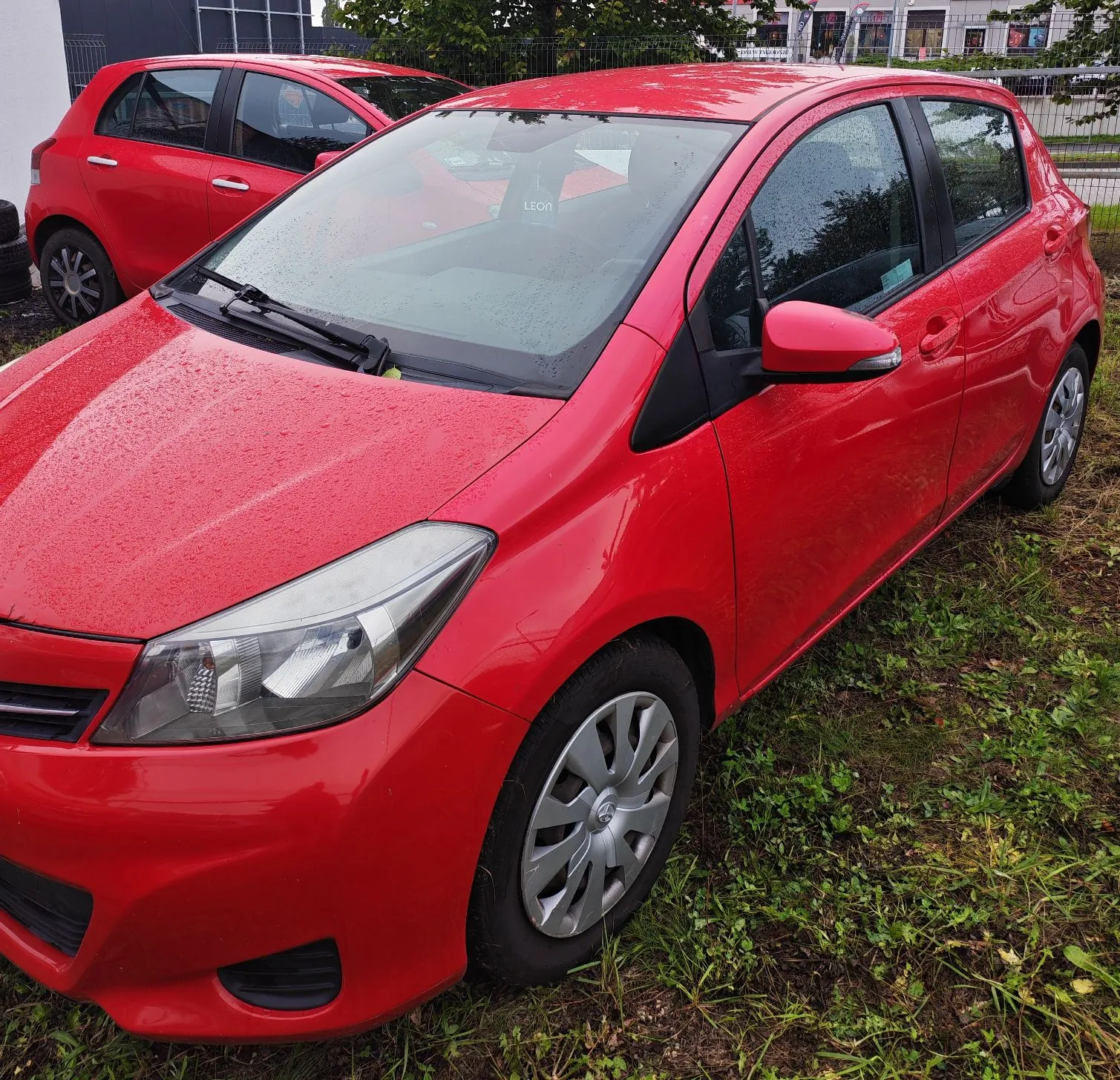 Czerwony samochód osobowy typu hatchback zaparkowany na trawie. Widoczny jest przód i prawy bok pojazdu. W tle znajduje się drugi, podobny czerwony samochód oraz fragment ogrodzenia i budynku.