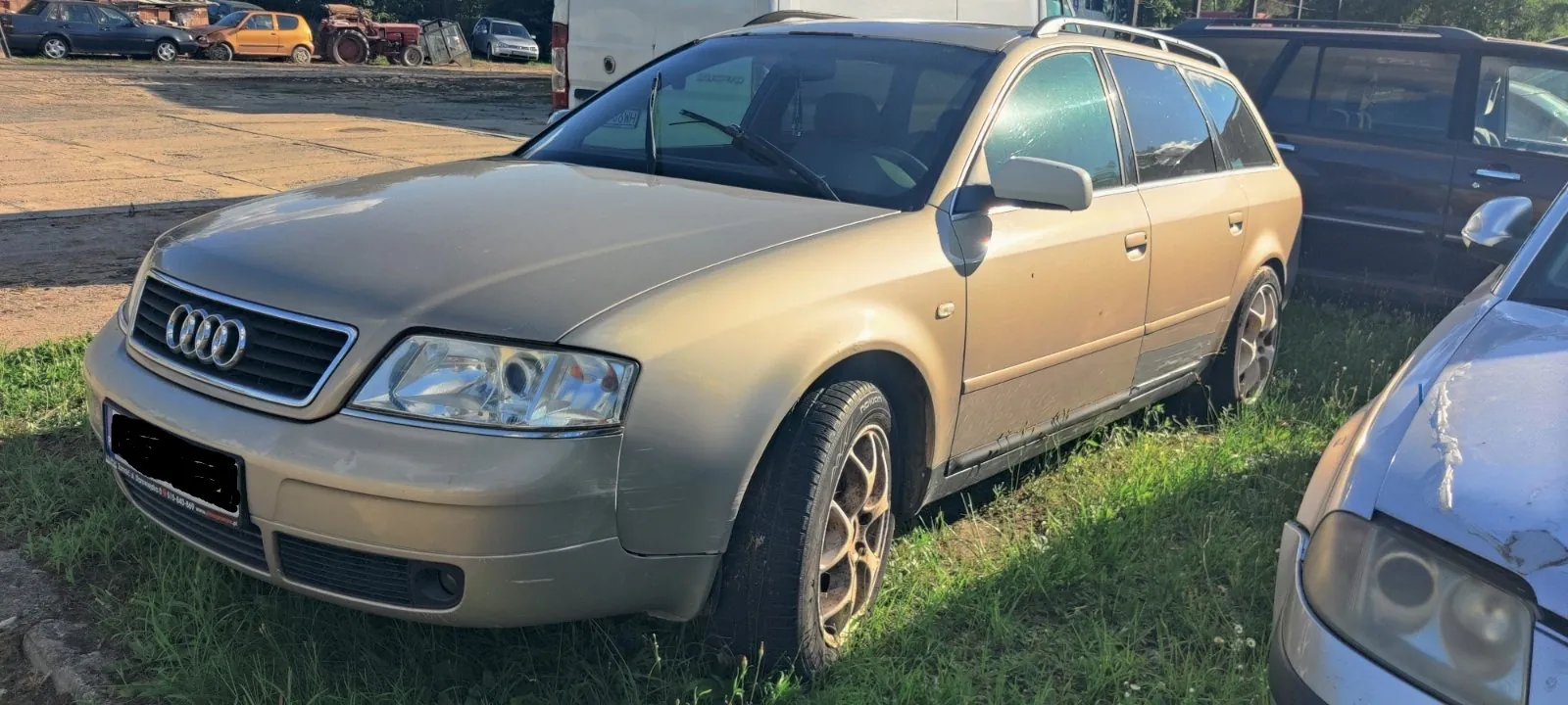 Złoty samochód marki Audi w nadwoziu kombi, zaparkowany na trawie obok innych pojazdów. Widoczny jest przód auta z charakterystycznym grillem i czterema pierścieniami logo Audi, reflektory oraz przednie prawe koło z brązową felgą. W tle znajdują się inne samochody i utwardzony plac.