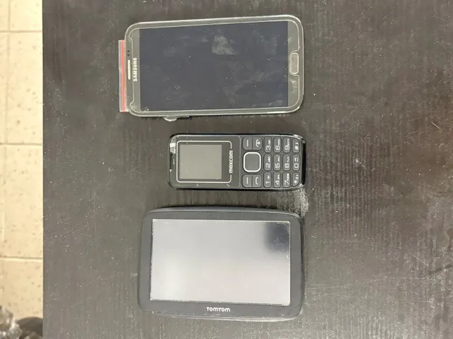 Trzy urządzenia elektroniczne ułożone pionowo na ciemnej, drewnianej powierzchni. Na górze znajduje się smartfon marki Samsung z czarnym ekranem, ułożony poziomo. W środku znajduje się mały, czarny telefon komórkowy z fizyczną klawiaturą i ekranem z napisem 'maxcom'. Na dole znajduje się urządzenie nawigacyjne marki TomTom z dużym, czarnym ekranem.
