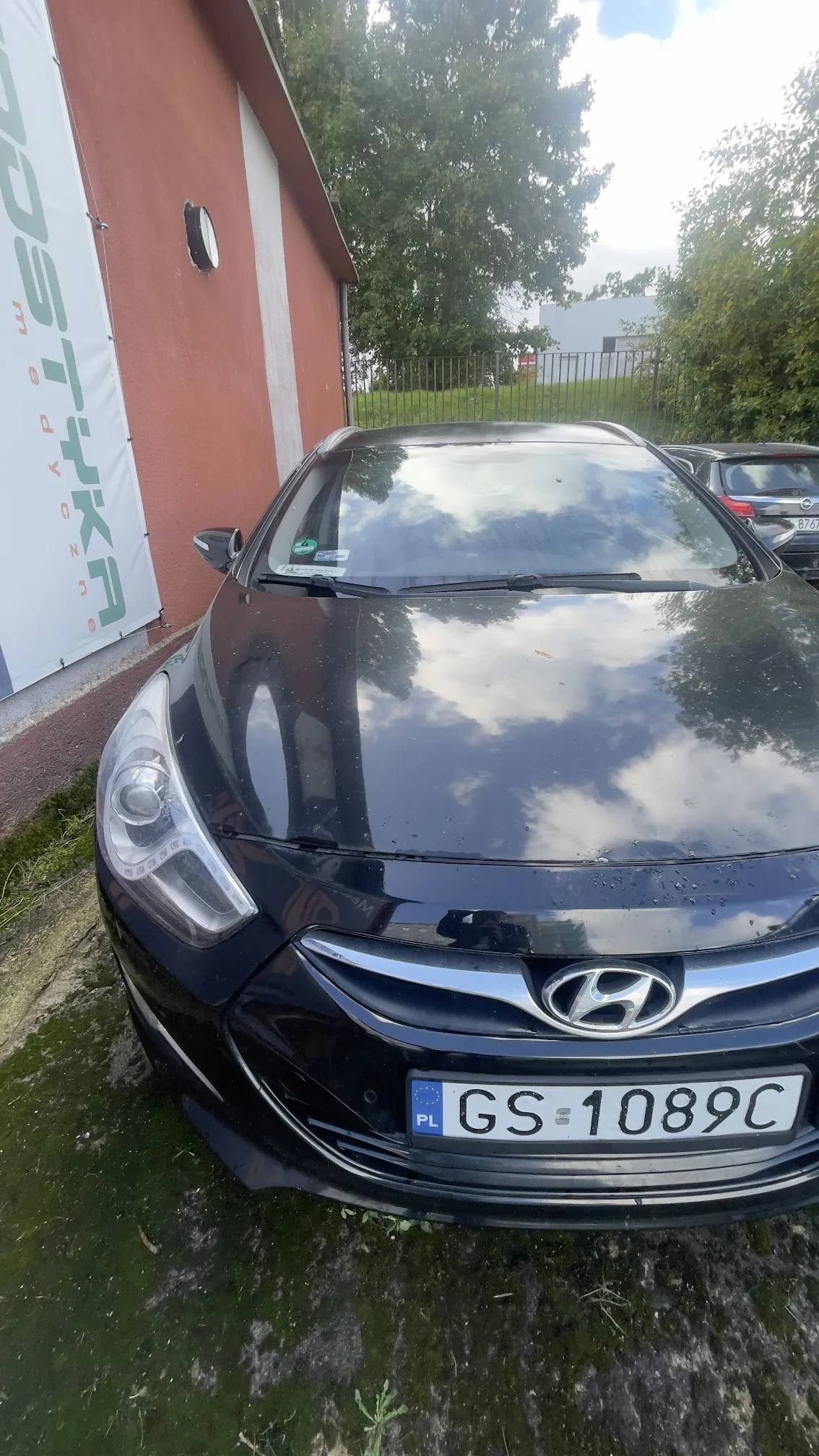 Czarny samochód marki Hyundai zaparkowany przed czerwonym budynkiem. Na masce samochodu odbijają się chmury. Pojazd posiada polską tablicę rejestracyjną o numerze GS 1089C. Po lewej stronie widoczny jest fragment białego baneru z napisem 'LOGISTYKA'.