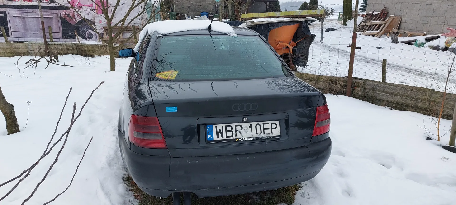 Czarny samochód marki Audi z tyłu, zaparkowany na śnieżnej powierzchni. Na dachu widoczny śnieg, a na szybie tylnej żółty znak. Tablica rejestracyjna PL WBR 10EP z naklejką X-CARwash. W tle siatka ogrodzeniowa, nagie drzewa, betonowa ściana, pomarańczowa maszyna przykryta czarną folią oraz budynek z muralami. Śnieg pokrywa całą okolicę, w tym ziemię i przedmioty w tle.