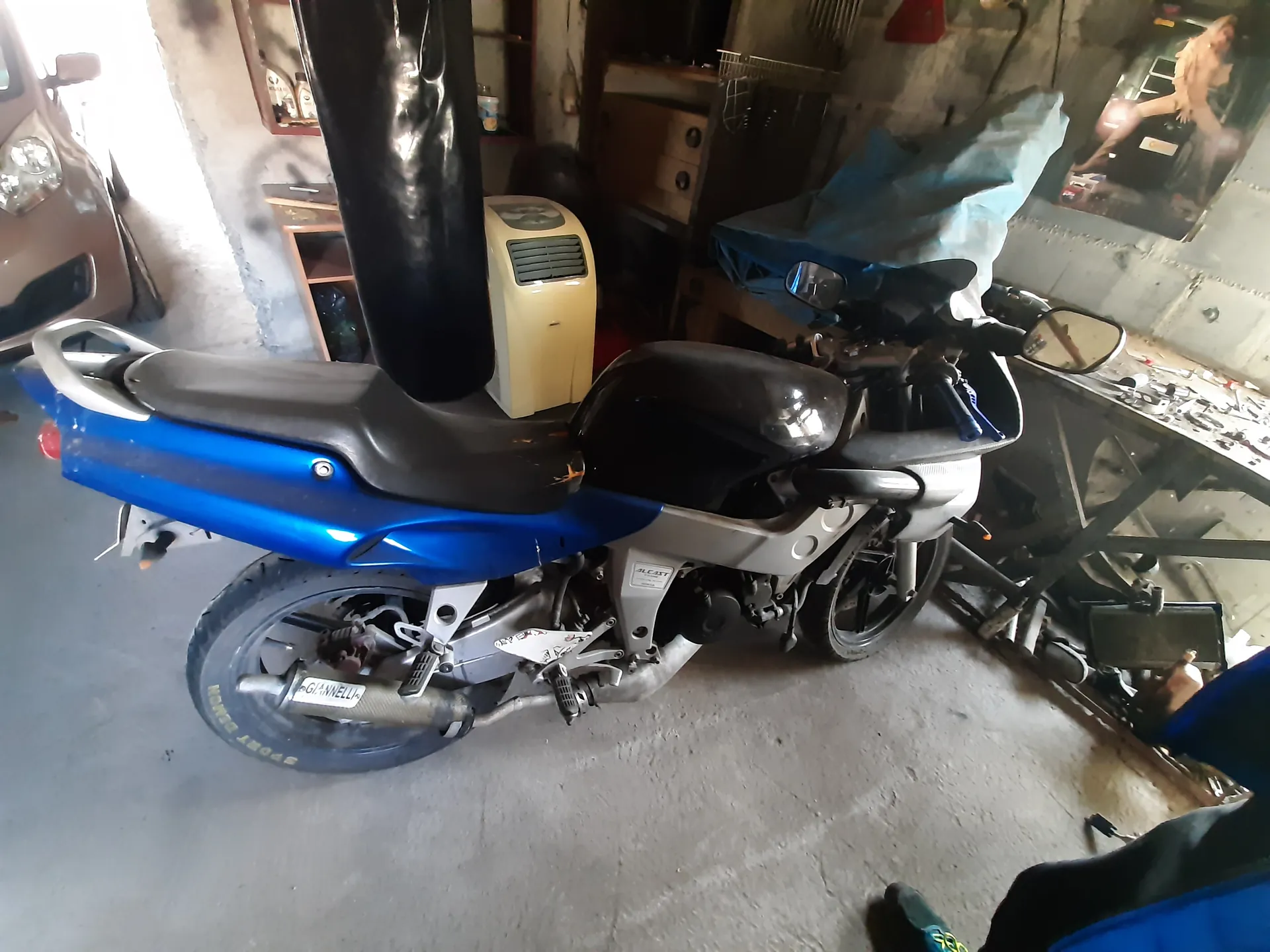 Niebiesko-czarny motocykl w garażu. Widoczny napis 'GIANNELLI' na wydechu oraz napis 'SPORT' na oponie tylnego koła. W tle częściowo widoczny samochód, żółty aparat, narzędzia na stole, niebieska folia zakrywająca przedmiot oraz plakat na ścianie. Podłoga z betonu.