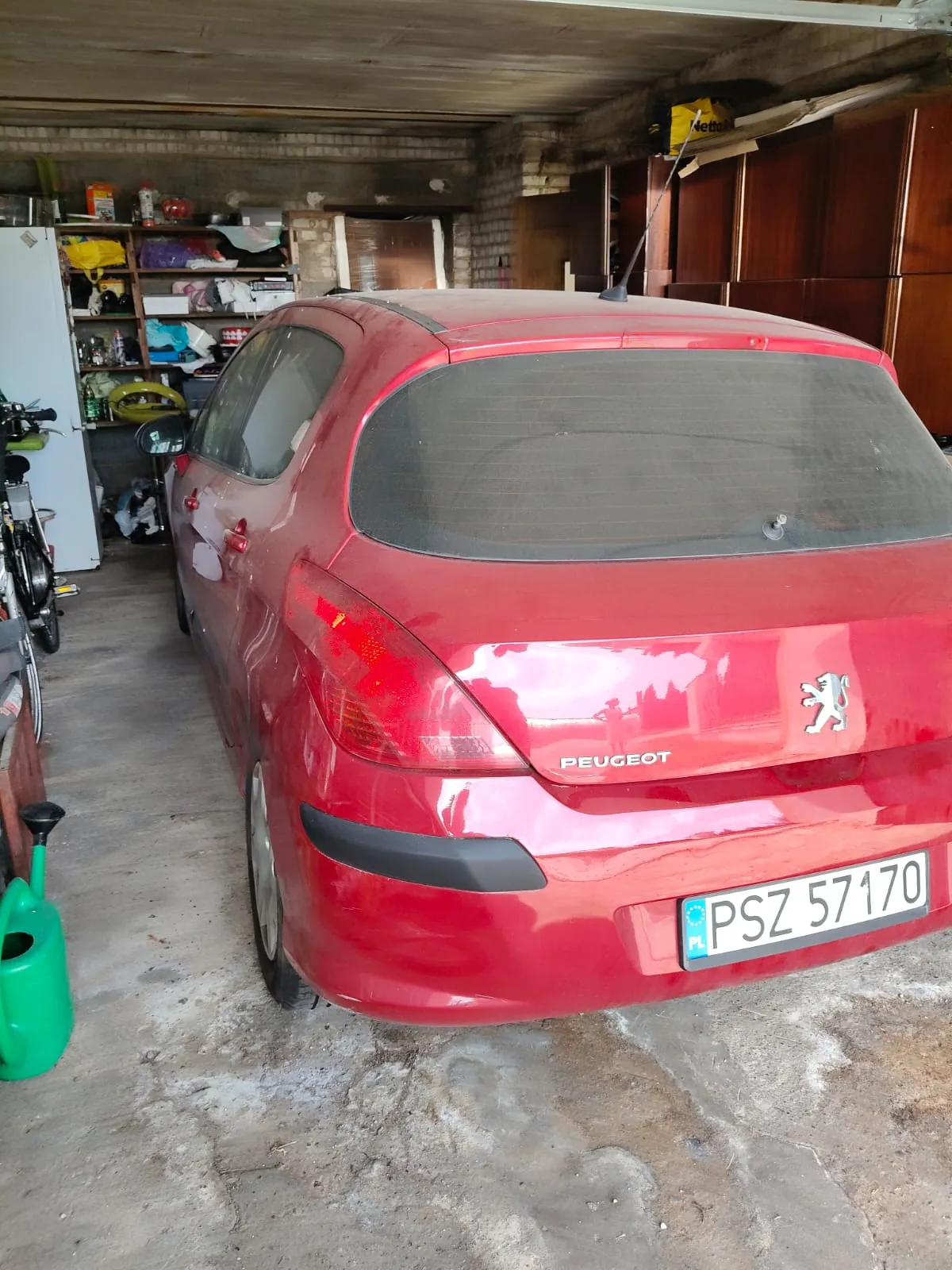 Tylny widok czerwonego samochodu marki Peugeot zaparkowanego w garażu. Na klapie bagażnika widnieje napis PEUGEOT oraz logo marki. Samochód ma polską tablicę rejestracyjną o numerze PSZ 57170. W tle widoczne są półki z różnymi przedmiotami oraz drewniane szafki.