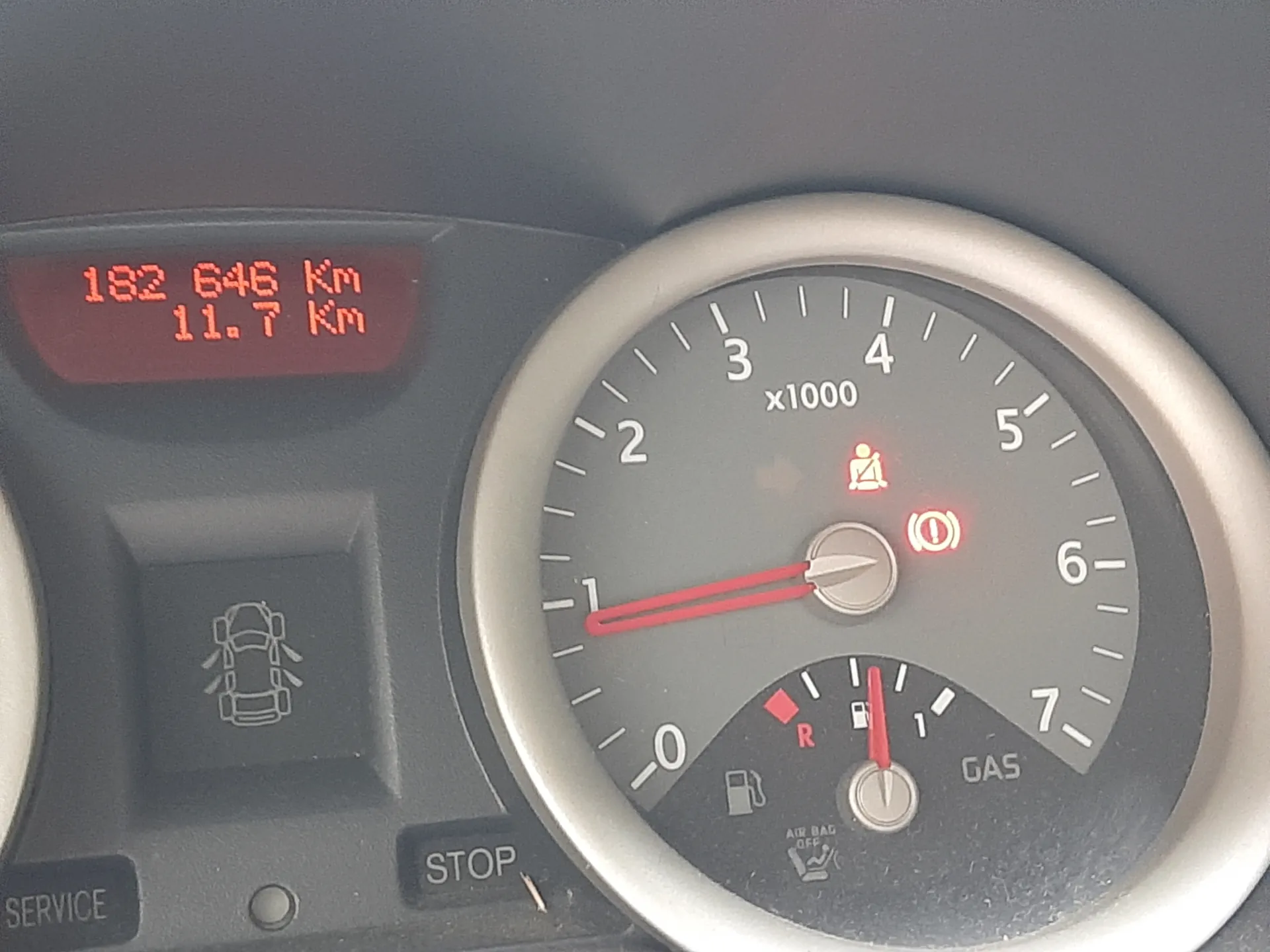 Zbliżenie na deski rozdzielcze samochodu. Po lewej stronie wyświetlacz cyfrowy z czerwonymi cyframi: 182 646 Km i 11.7 Km. Poniżej znajduje się przycisk z ikoną samochodu oraz napisem SERVICE. W centrum tachometr z zakresem 0-7 (x1000), czerwonym wskazówką na pozycji 1 i dwoma aktywnymi lampkami ostrzegawczymi: pas bezpieczeństwa i symbol silnika. Poniżej tachometru wskaźnik paliwa z czerwoną wskazówką, napisem GAS oraz literą R. W dolnej części widoczny przycisk STOP i napis AIR BAG OFF.