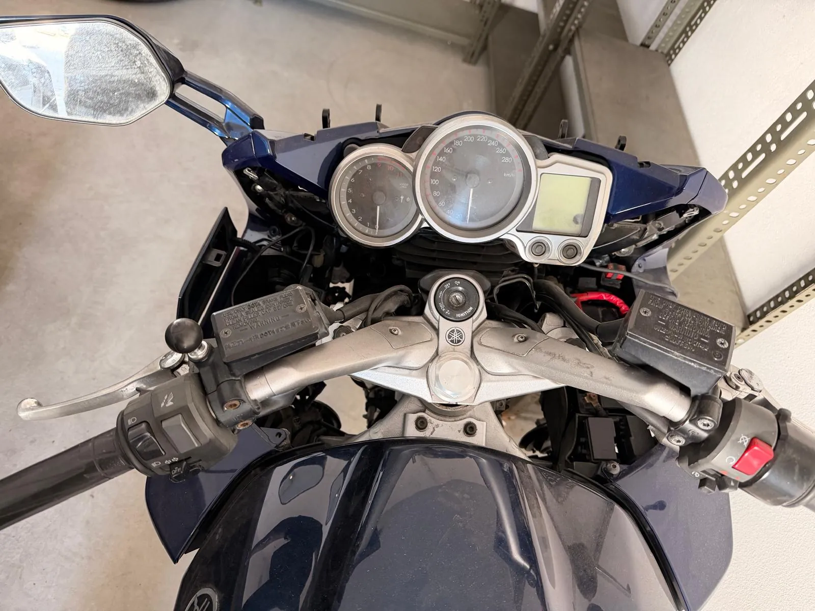 Widok z perspektywy kierowcy na kokpit ciemnoniebieskiego motocykla. Widoczny jest licznik prędkości, obrotomierz, kierownica z manetkami oraz lusterko wsteczne po lewej stronie. Obraz jest zorientowany pionowo, podczas gdy motocykl jest ustawiony poziomo.