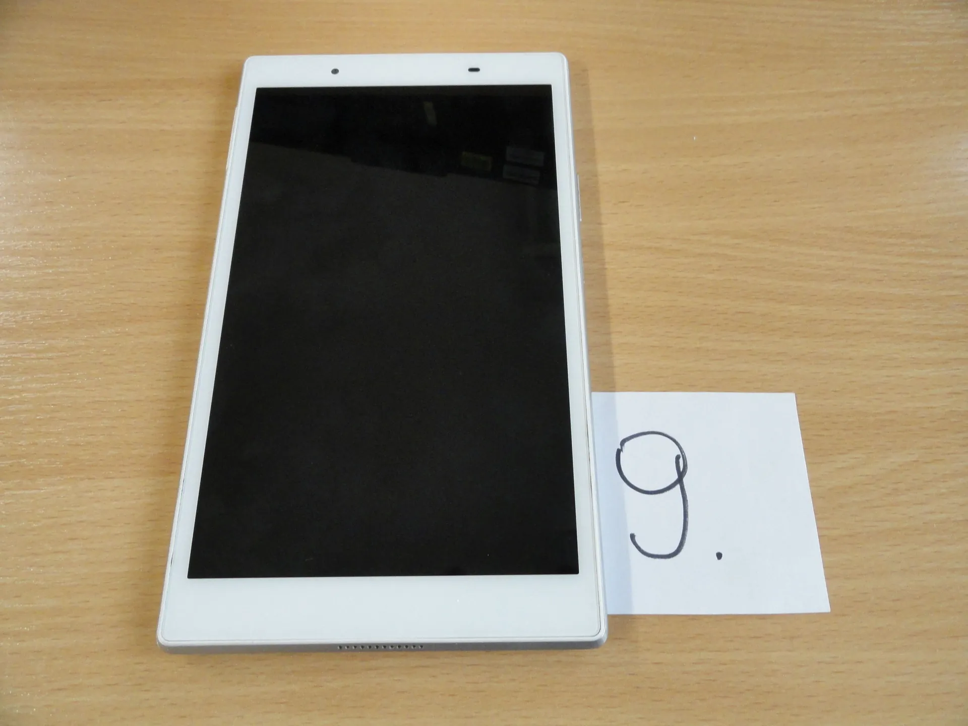 Biały tablet z czarnym ekranem leżący na drewnianym blacie. Obok tabletu znajduje się biały arkusz papieru z ręcznie napisaną cyfrą 9. i kropką.