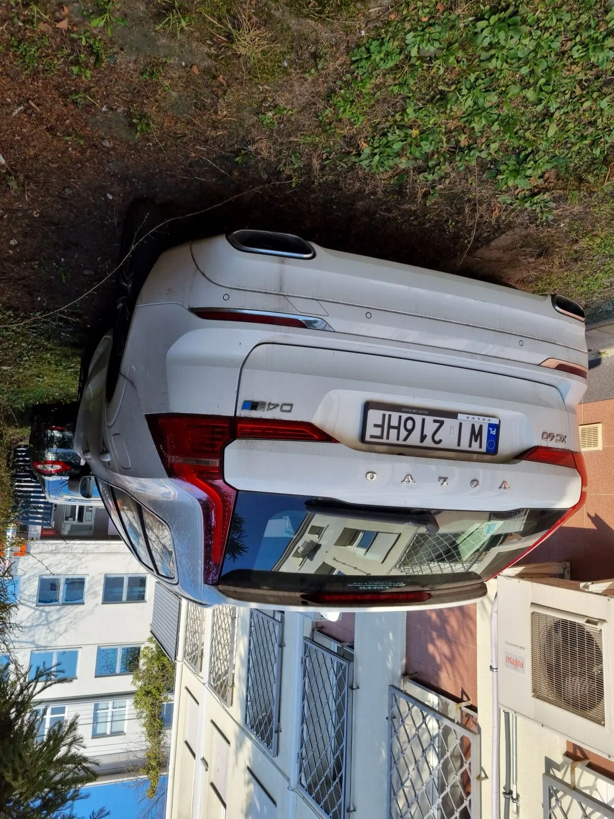 Srebrny samochód Volvo XC60 z tablicą rejestracyjną WI 216HF zaparkowany przy budynku z białą elewacją, oknami z metalowymi kratami i jednostką klimatyzacyjną. W tle widoczne są inne budynki, drzewa oraz trawnik. Na tylnym zderzaku samochodu widoczny napis D4 i XC60.