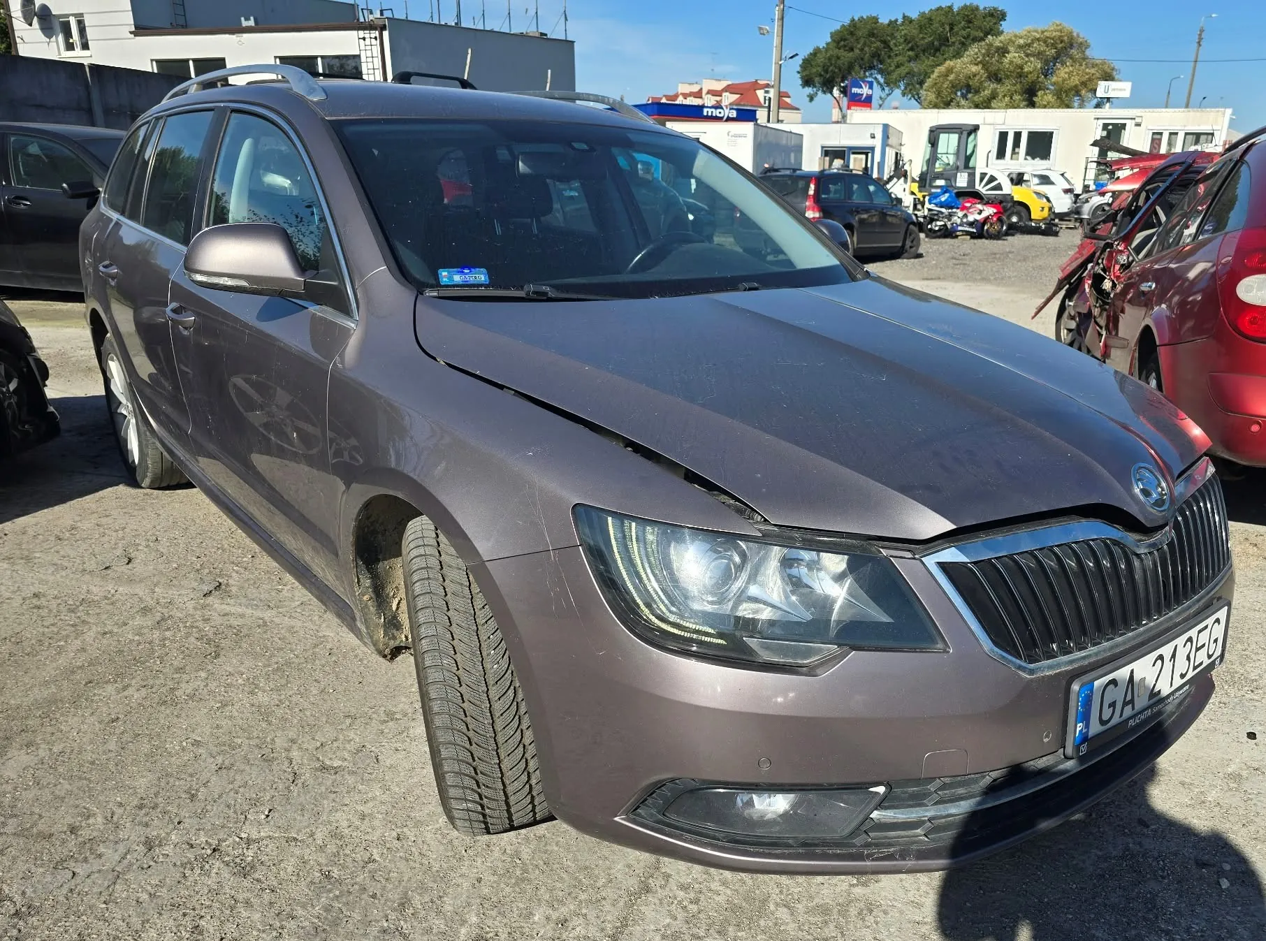 Brązowy samochód marki Skoda w nadwoziu kombi, zaparkowany na betonowym placu. Pojazd posiada uszkodzenia karoserii w przedniej części: maska jest lekko podniesiona i niedomknięta, a przedni zderzak jest pęknięty i zarysowany po prawej stronie. Samochód ma tablicę rejestracyjną o numerze GA 213EG. W tle widoczne są inne samochody, budynki przemysłowe oraz niebieski szyld z napisem 'moja'.