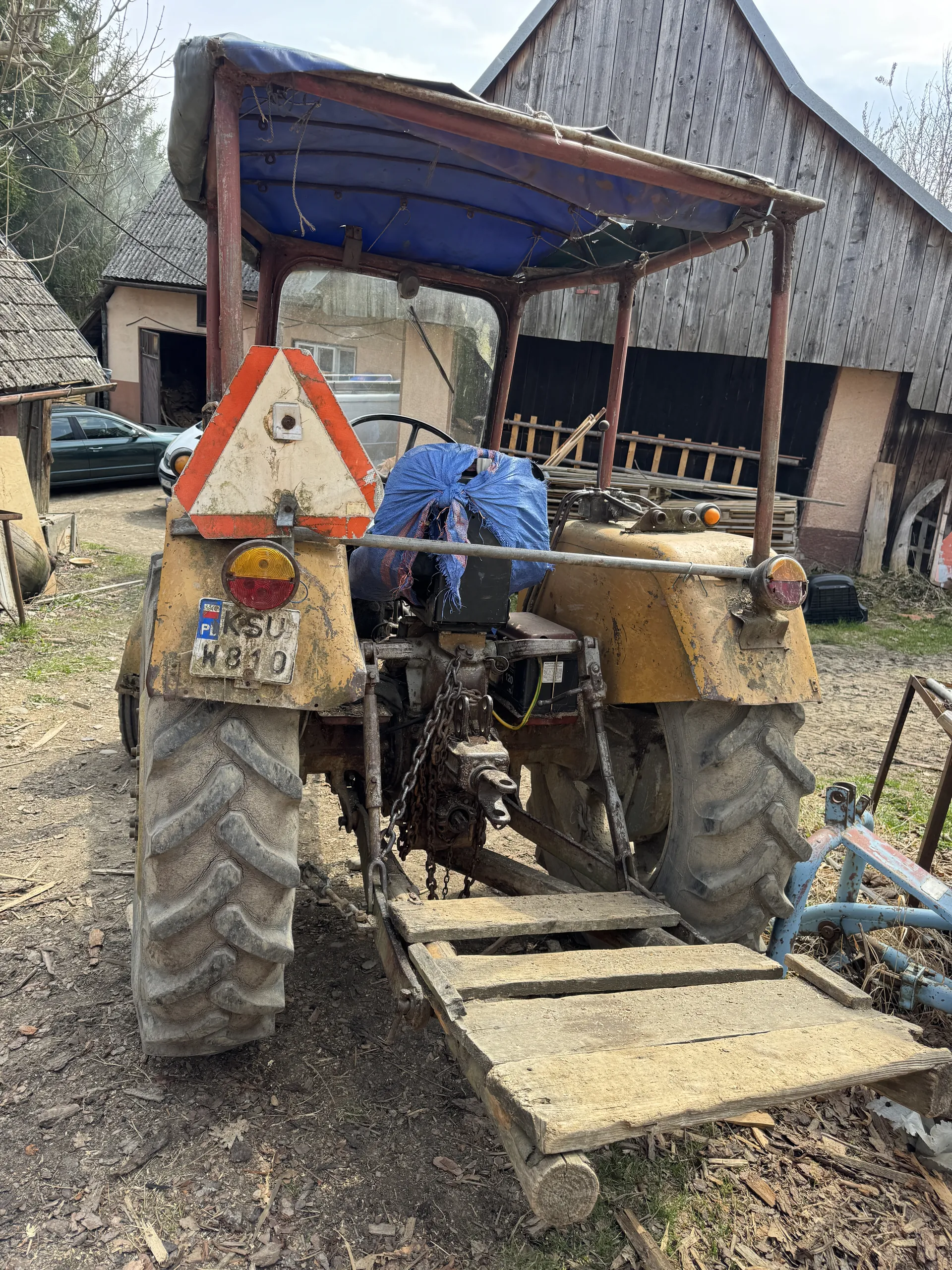 Stary żółty traktor z dużymi, brudnymi oponami. Na siedzeniu znajduje się niebieska tkanina. Przednia część traktora posiada trójkątny znak ostrzegawczy pomarańczowo-biały oraz tablicę rejestracyjną "KSU W810" z małą naklejką flagi Polski. W tle widoczne są drewniane budynki, drabina, samochód oraz narzędzia. Podłoże pokryte jest ziemią i wiórami drewnianymi.