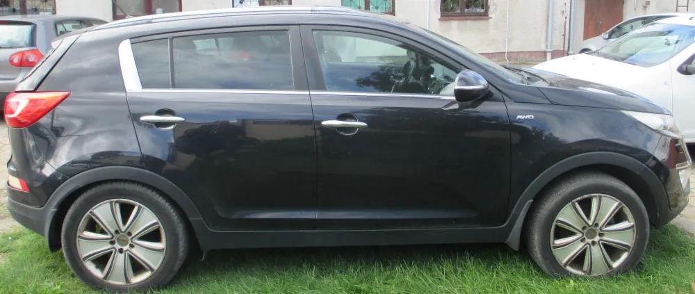 Czarny samochód typu SUV zaparkowany bokiem na trawie. Pojazd posiada srebrne klamki, srebrne wykończenie wokół okien oraz felgi aluminiowe z pięcioma szerokimi ramionami. Na przednim błotniku widoczny jest mały napis 'AWD'. W tle widoczne są fragmenty innych samochodów oraz jasna ściana budynku.