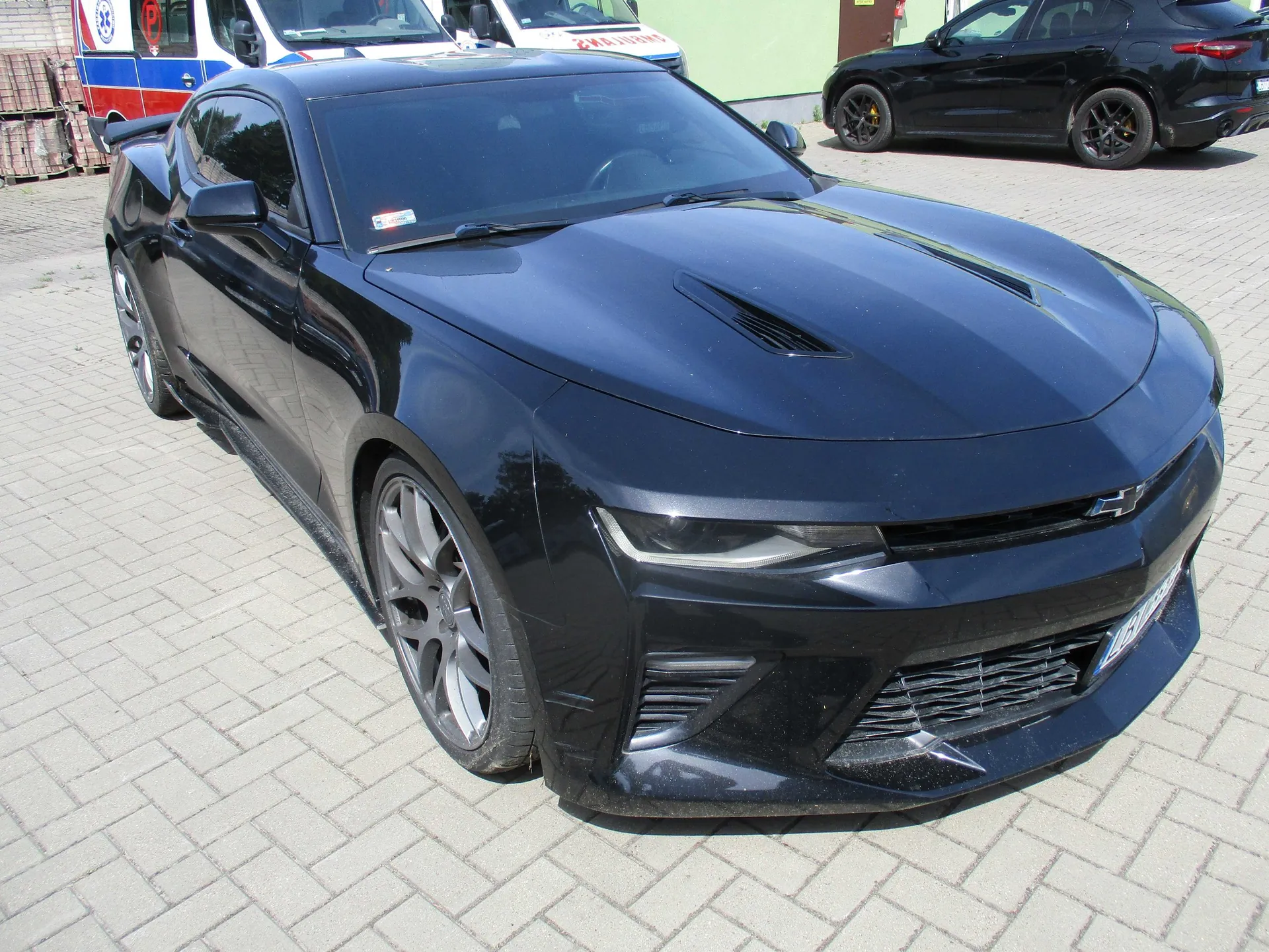 Czarny samochód sportowy Chevrolet Camaro zaparkowany na kostce brukowej. Auto jest widoczne z przodu i z boku. W tle widać fragmenty białych pojazdów służbowych oraz czarny samochód osobowy.