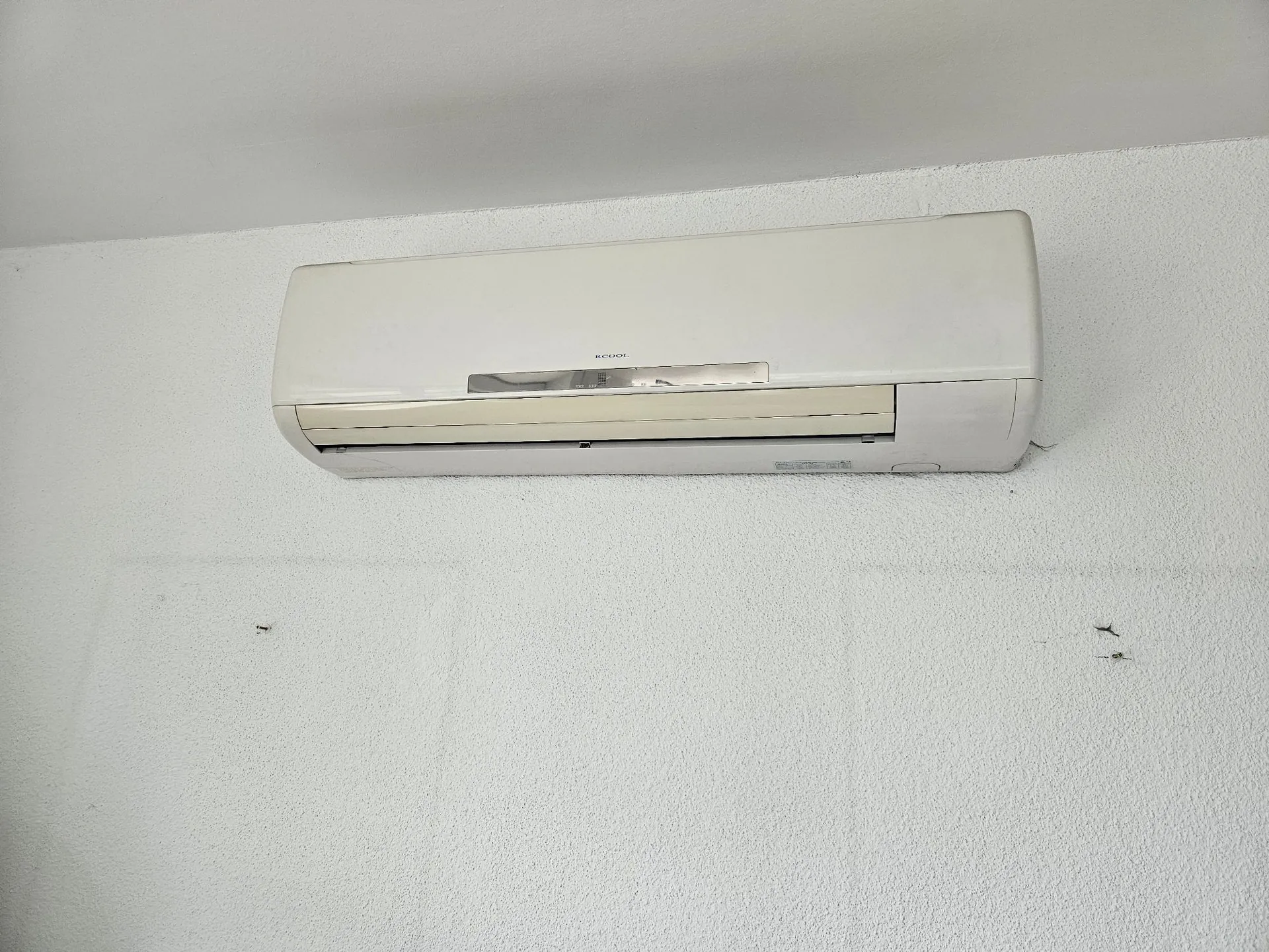 Biała jednostka wewnętrzna klimatyzatora zamontowana wysoko na białej, chropowatej ścianie pod sufitem. Na obudowie urządzenia widnieje napis 'RCOOL'.