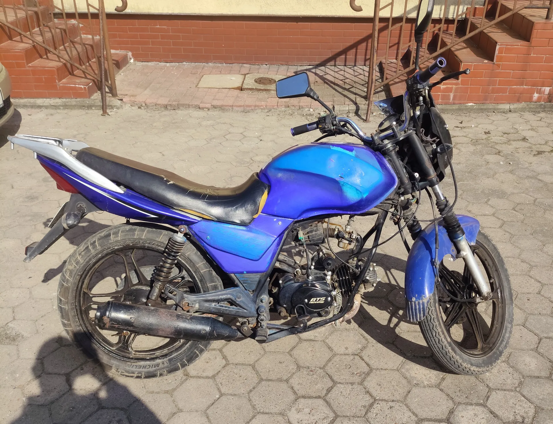Niebieski motocykl z czarnym siedzeniem zaparkowany na chodniku z sześciokątnymi płytami. Widoczne są elementy konstrukcyjne, w tym silnik z napisem BTS, lusterko boczne, kierownica oraz tylne i przednie koło. W tle ceglana ściana budynku, schody z metalowymi poręczami oraz fragment innego pojazdu po lewej stronie. Oświetlenie słoneczne rzuca cienie na powierzchnię.