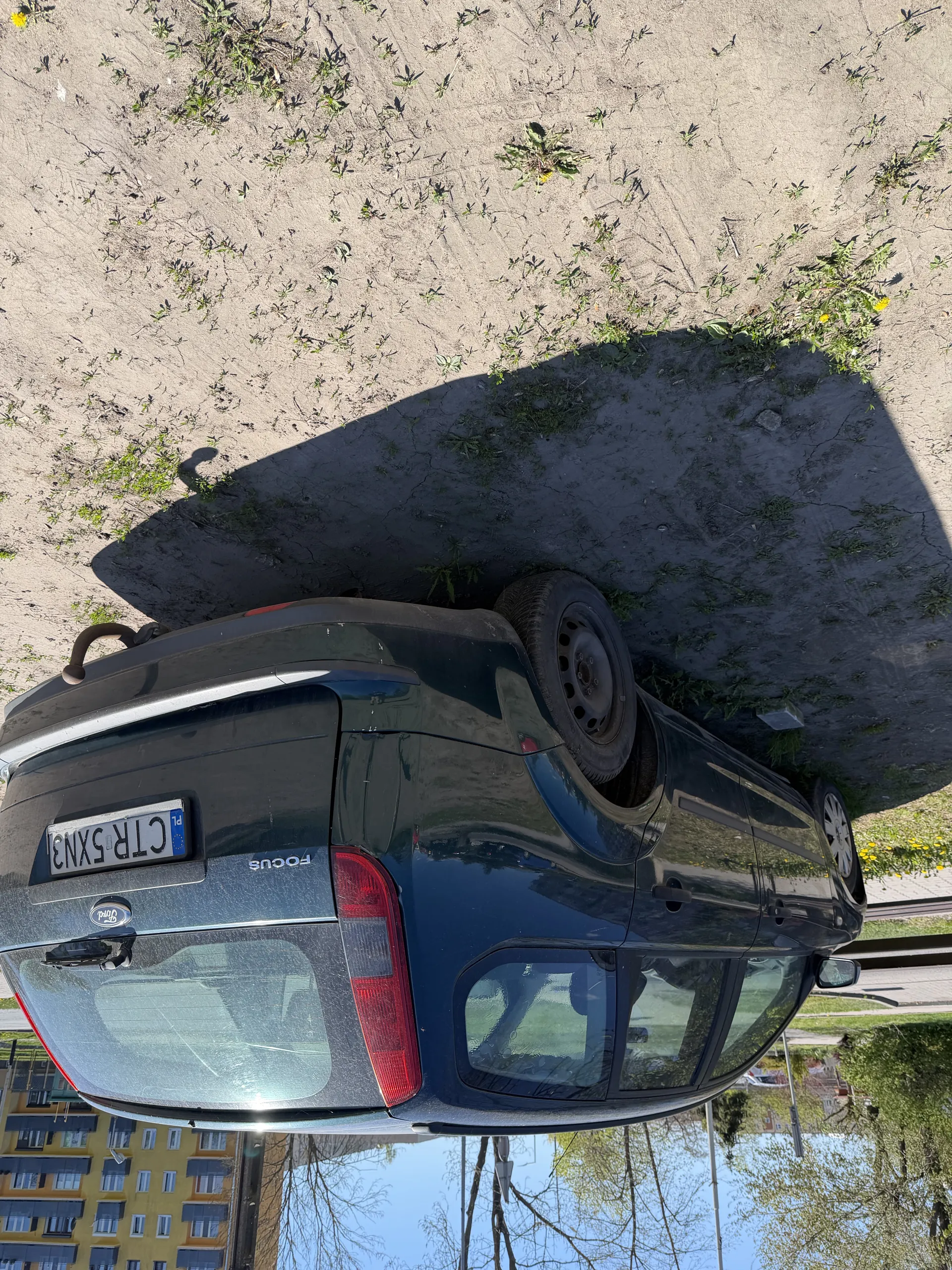 Ciemny samochód Ford Focus z tablicą rejestracyjną CTR 5XN3 zaparkowany na piaszczystej powierzchni z rozproszonymi roślinami. W tle widoczny jest żółto-szary budynek wielorodzinny, drzewa oraz jasne niebo. Samochód zajmuje centralną część obrazu, a jego tył jest skierowany w górę.