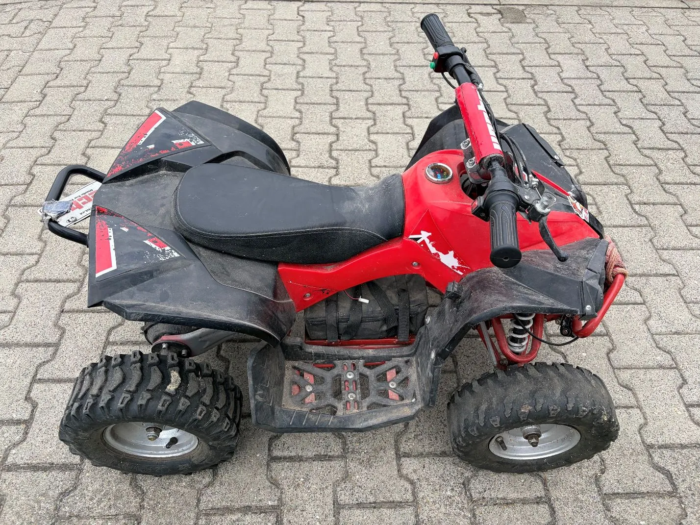 Czerwono-czarny quad stojący na szarym, kostkowym podłożu. Pojazd posiada szerokie, terenowe opony, czarne błotniki oraz czarne siedzenie. Na czerwonych elementach nadwozia widoczne są białe i czarne grafiki.