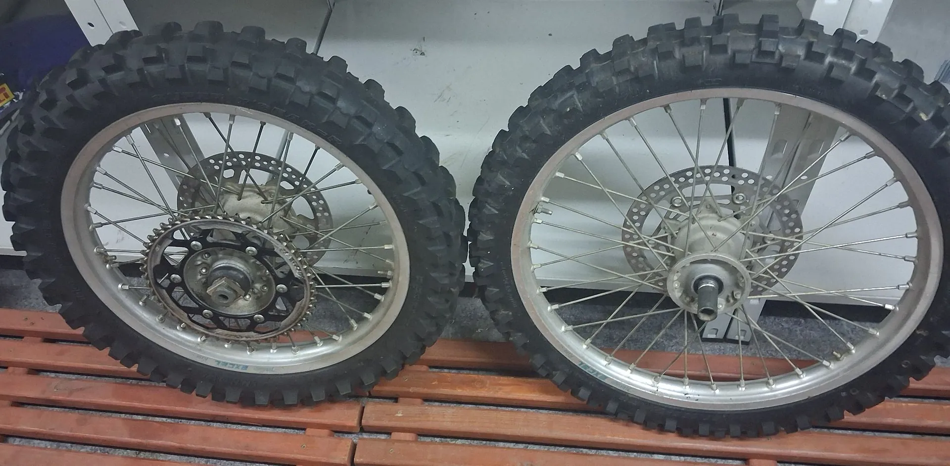 Dwa koła od motocykla terenowego z grubymi, ząbkowanymi oponami typu kostka, ustawione obok siebie na drewnianej palecie. Lewe koło posiada zamontowaną zębatkę napędową, oba koła mają szprychowe obręcze i tarcze hamulcowe.