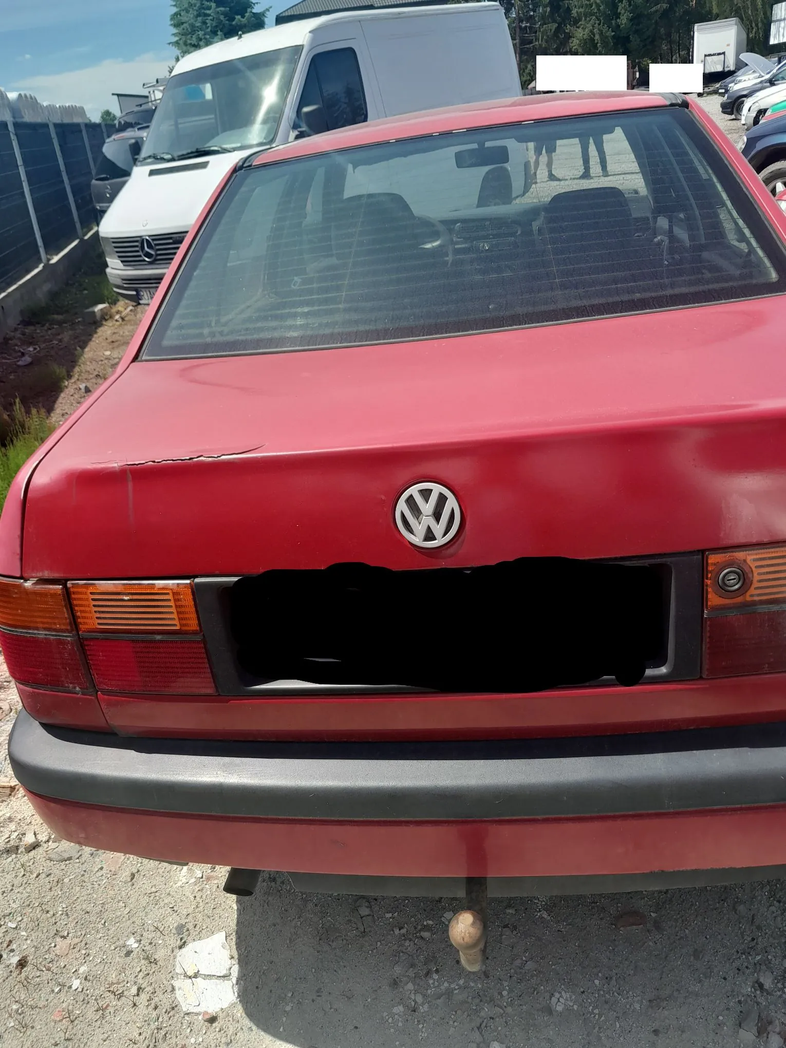 Tylny widok czerwonego samochodu marki Volkswagen. Na klapie bagażnika widoczne jest logo marki oraz wgniecenie w lewej górnej części. Tablica rejestracyjna jest zakryta czarnym polem. Samochód posiada czarny zderzak i hak holowniczy. W tle widoczny jest biały furgon marki Mercedes-Benz oraz inne zaparkowane pojazdy.