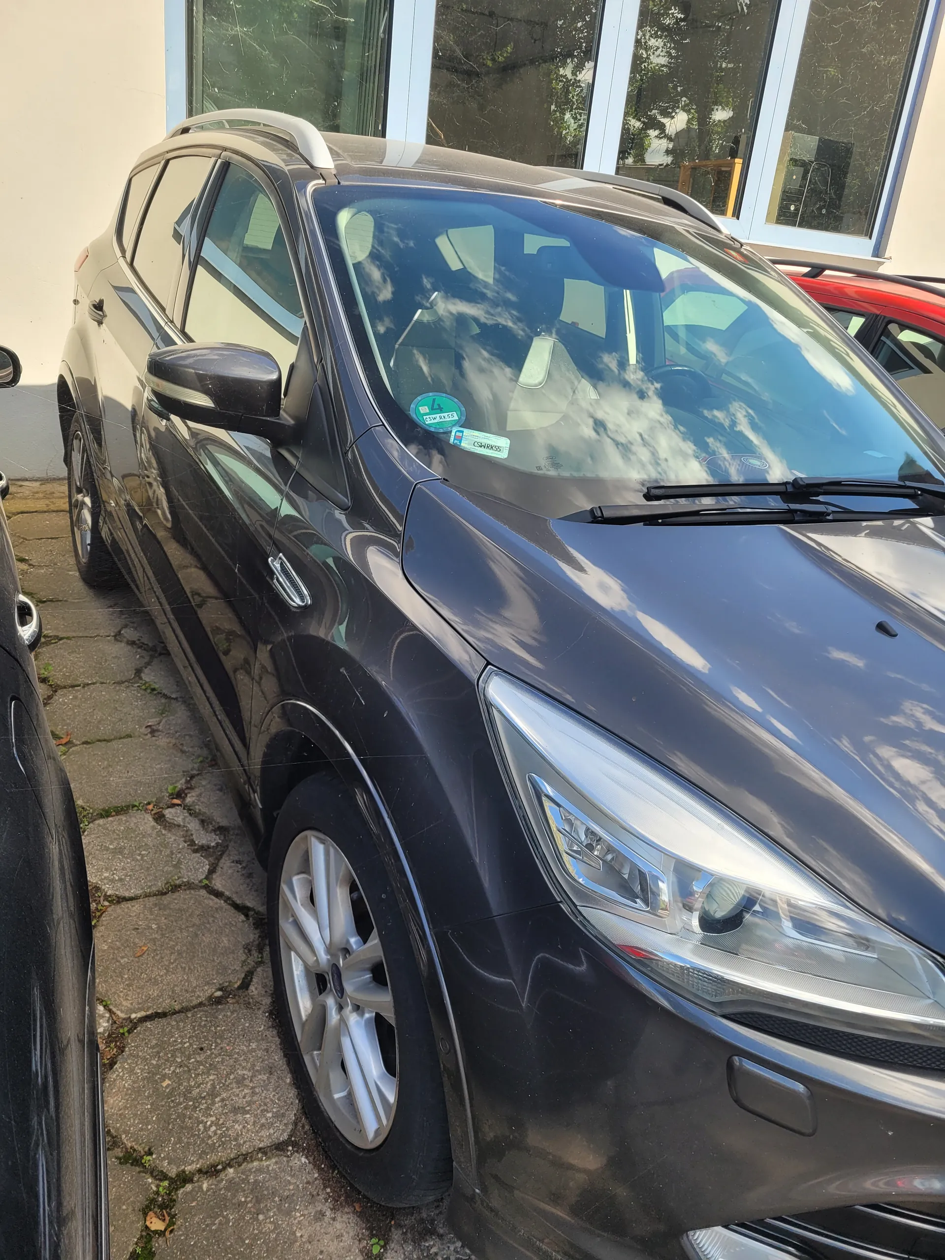 Ciemnoszary samochód widziany od przodu z widocznym przednim reflektorem, przednim kołem z srebrnymi felgami i przednią szybą. Na przedniej szybie znajdują się naklejki: zielona z cyfrą '4' oraz biała z napisem '2024'. W tle budynki z dużymi oknami i fragment czerwonego samochodu. Samochód zaparkowany na brukowanej powierzchni.