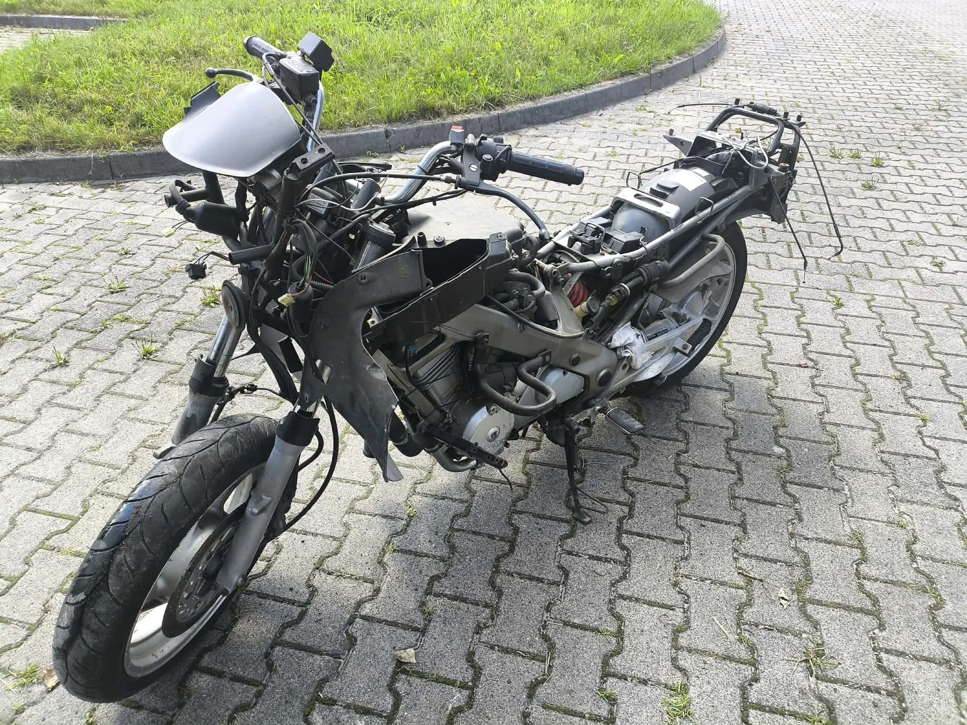 Zdjęcie uszkodzonego motocykla w kolorze szarym, który jest w stanie rozbiórki lub po wypadku. Brakuje wielu elementów nadwozia, siedzenia oraz tylnego koła. Motocykl stoi na kostce brukowej obok trawnika.