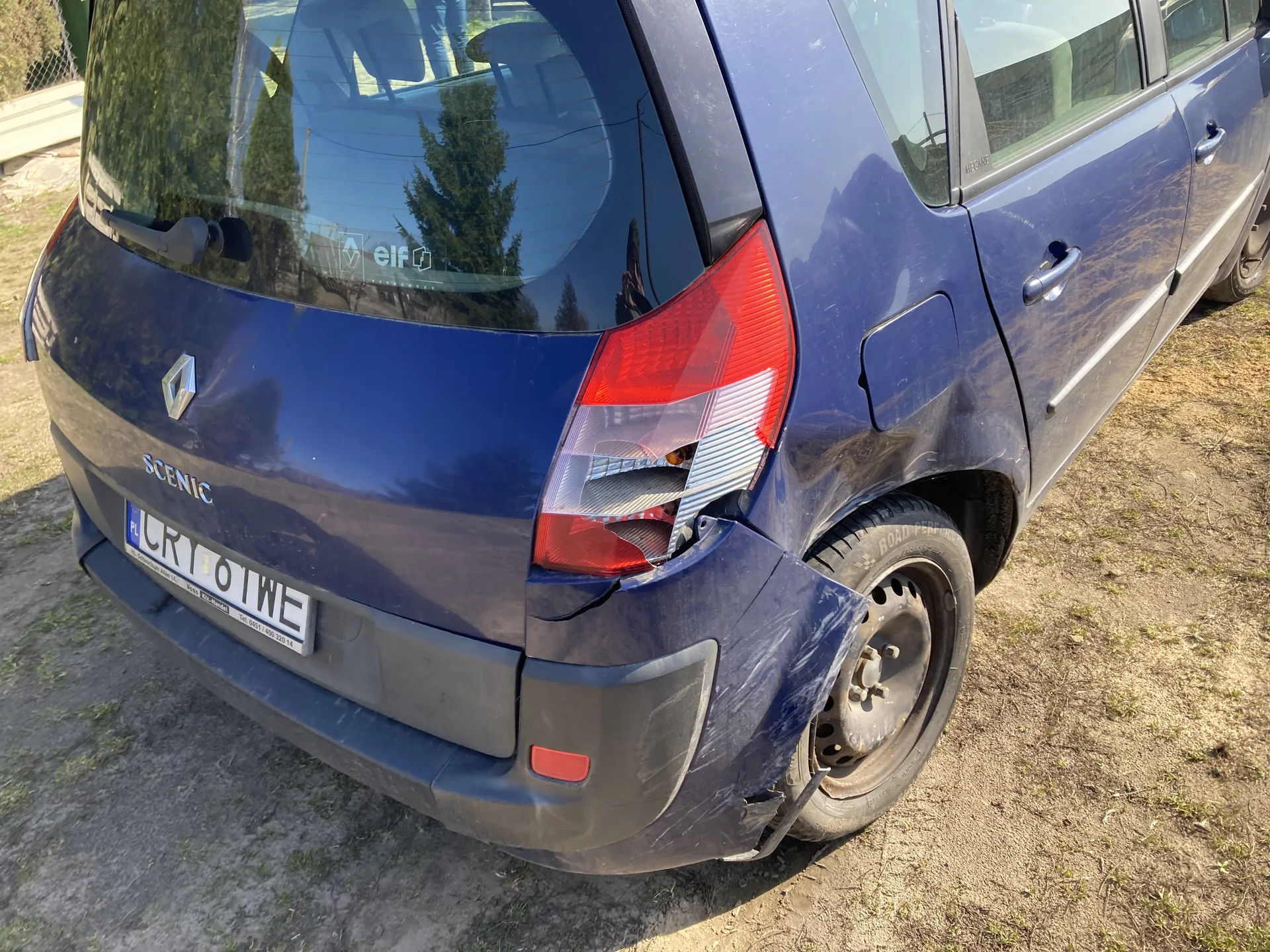 Niebieski samochód Renault Scenic z widocznym uszkodzeniem tylnego zderzaka i prawego tylnego światła. Na tylnej szybie naklejka z napisem 'elf'. Tablica rejestracyjna z polskim kodem 'PL' i numerem 'CRY 6 TWE'. Samochód zaparkowany na podwórku z suchą trawą i ziemią, w tle drzewa i ogrodzenie. Widoczne refleksy na szybie oraz logo marki na bagażniku.