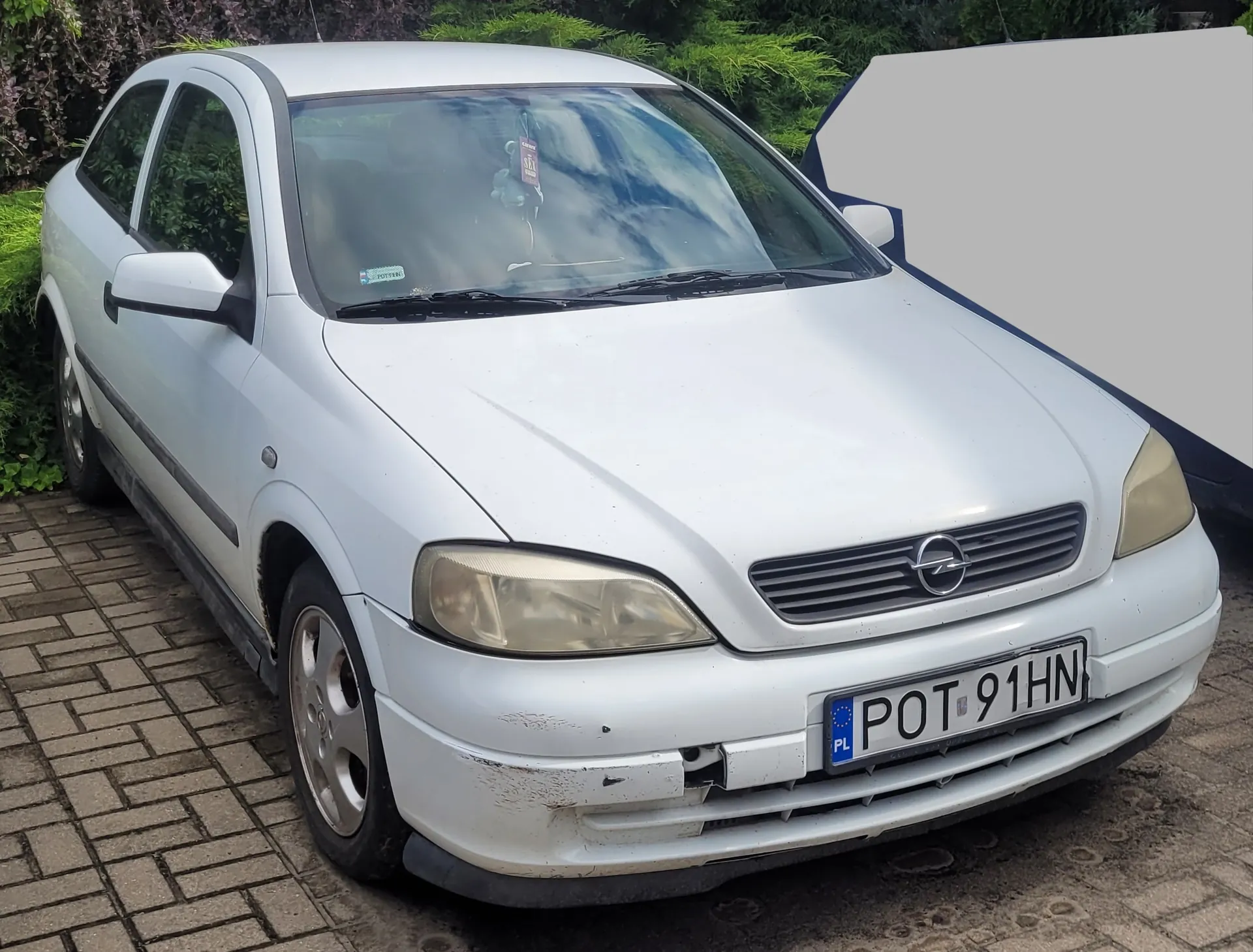 Biały samochód osobowy marki Opel zaparkowany na kostce brukowej. Pojazd jest widoczny z przodu i z boku. Na przednim zderzaku widoczne są uszkodzenia i zarysowania. Samochód posiada polską tablicę rejestracyjną o numerze POT 91HN.