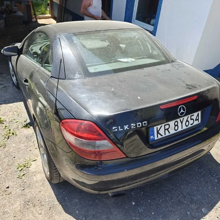 Czarny samochód Mercedes-Benz SLK 200 z tablicą rejestracyjną KR 8Y654 (PL) zaparkowany na żwirowym podjeździe. Widok od tyłu, z widocznym logo marki, napisem SLK 200 oraz czerwonymi światłami tylnymi. W tle budynek z białymi i niebieskimi ścianami oraz osoba w białej koszulce stojąca w drzwiach. Samochód pokryty jest kurzem, a na podjeździe rosną pojedyncze rośliny.