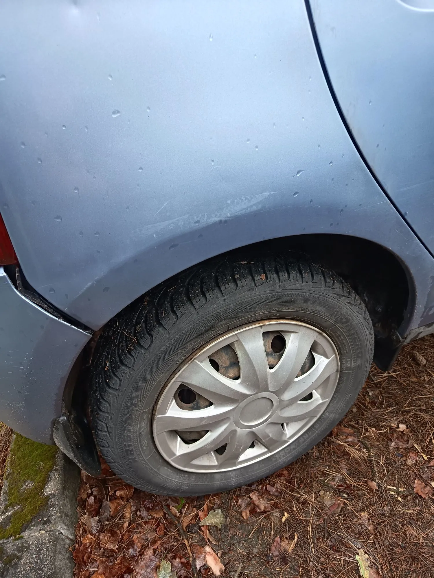 Zbliżenie na tylne koło samochodu w kolorze jasnoniebieskim. Na nadwoziu, powyżej nadkola, widoczne są zarysowania i drobne uszkodzenia lakieru. Koło posiada srebrną, plastikową osłonę felgi. Samochód stoi na podłożu pokrytym brązowymi, suchymi liśćmi i igliwiem.