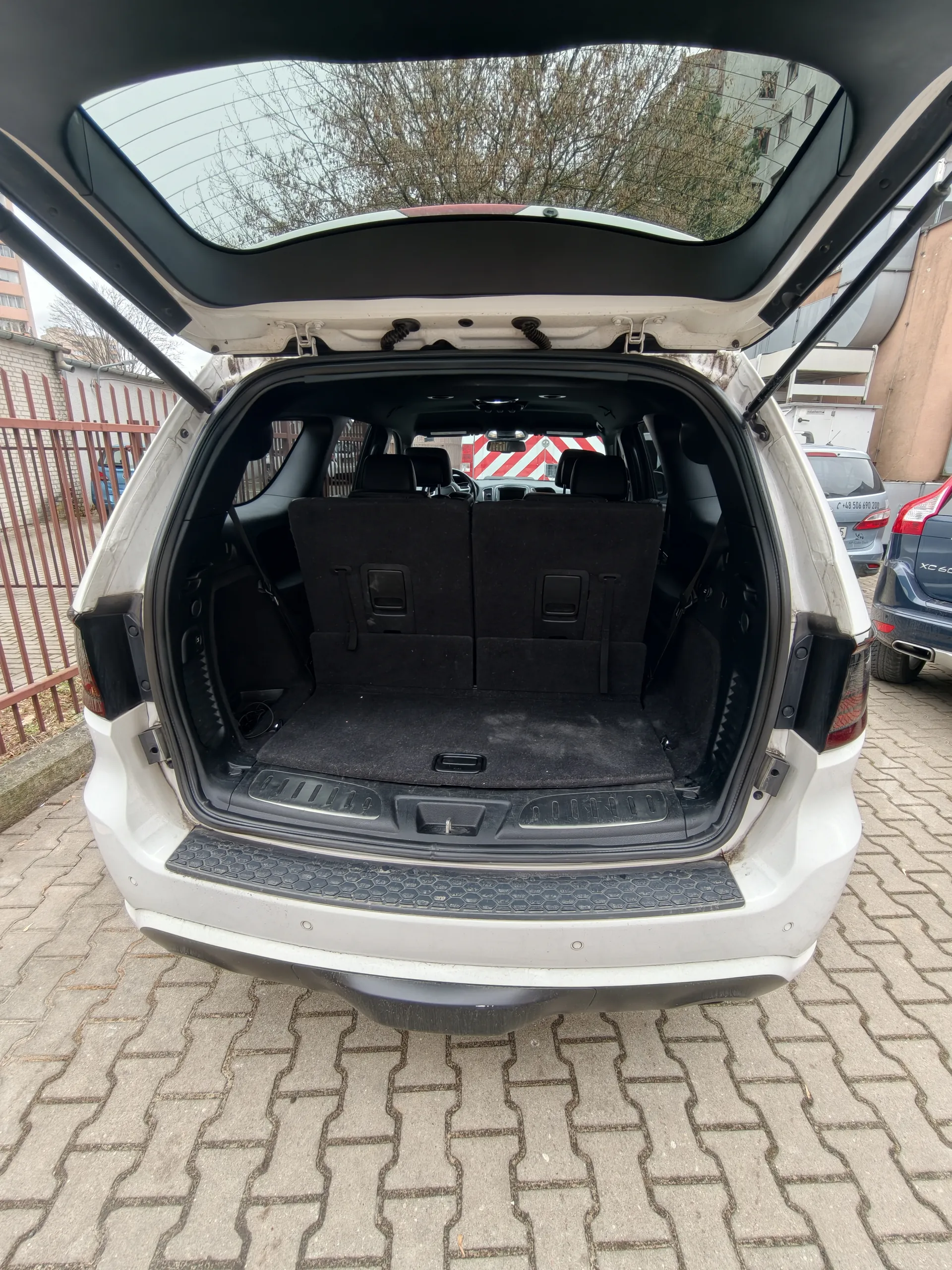Biały SUV z otwartym bagażnikiem, widok na pustą przestrzeń ładunkową i czarne siedzenia. Samochód zaparkowany na brukowanej powierzchni. W tle czerwone ogrodzenie, budynki, inne pojazdy (w tym samochód z widocznym napisem XC60) oraz drzewa bez liści. Niebo zachmurzone.