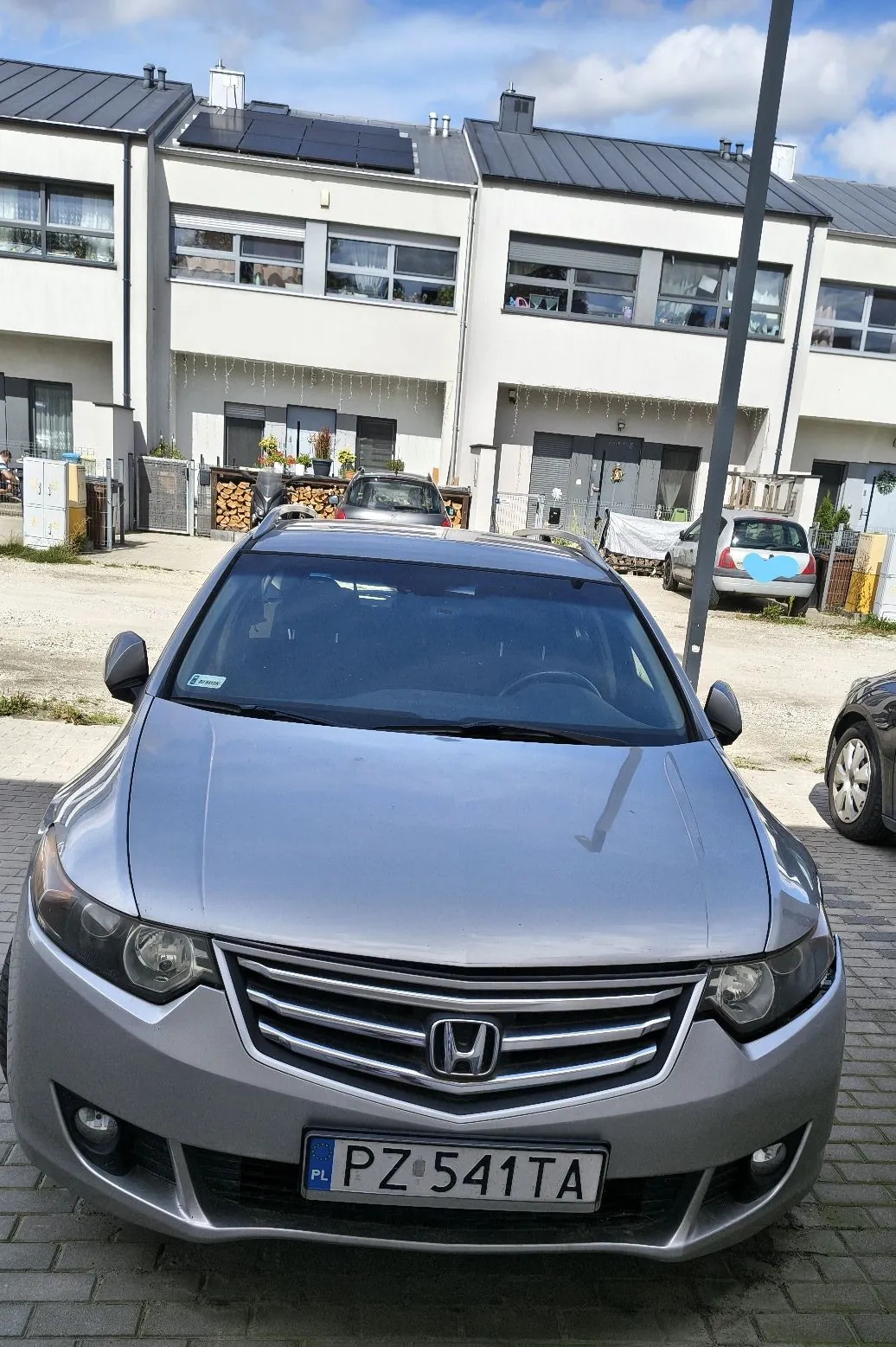 Srebrny samochód marki Honda zaparkowany przodem do obiektywu na kostce brukowej. Na samochodzie widoczna jest polska tablica rejestracyjna o numerze PZ 541TA. W tle znajdują się białe, nowoczesne domy szeregowe z panelami fotowoltaicznymi na dachach oraz inne zaparkowane samochody.