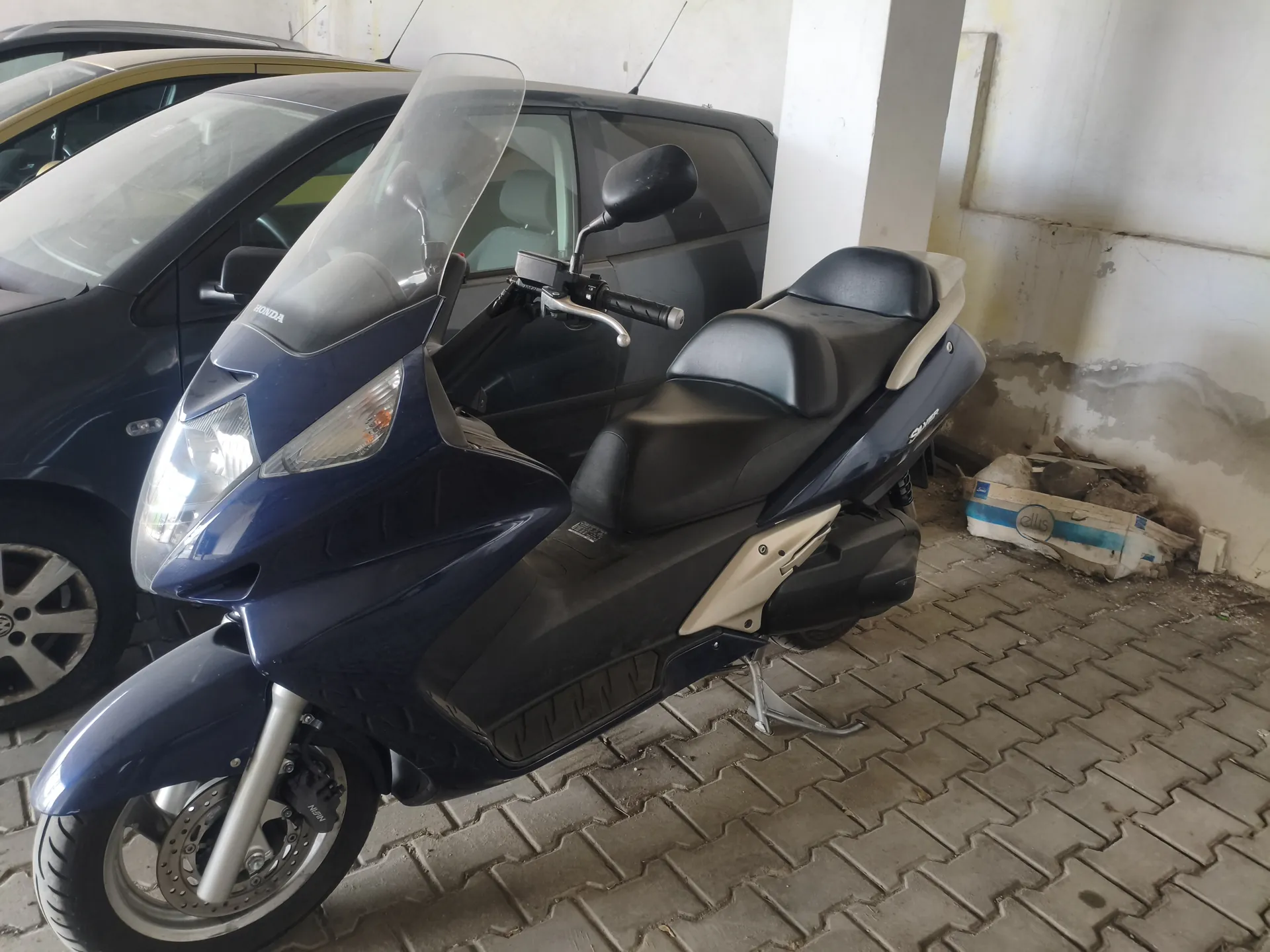 Niebieski skuter marki Honda z czarnym siedzeniem i beżowymi akcentami, wyposażony w przednią szybę i lusterka. Skuter zaparkowany na brukowanej powierzchni w pomieszczeniu z betonowymi ścianami, częściowo uszkodzonymi. W tle widoczny czarny samochód osobowy oraz pudełko z napisem 'ellis' na podłodze.