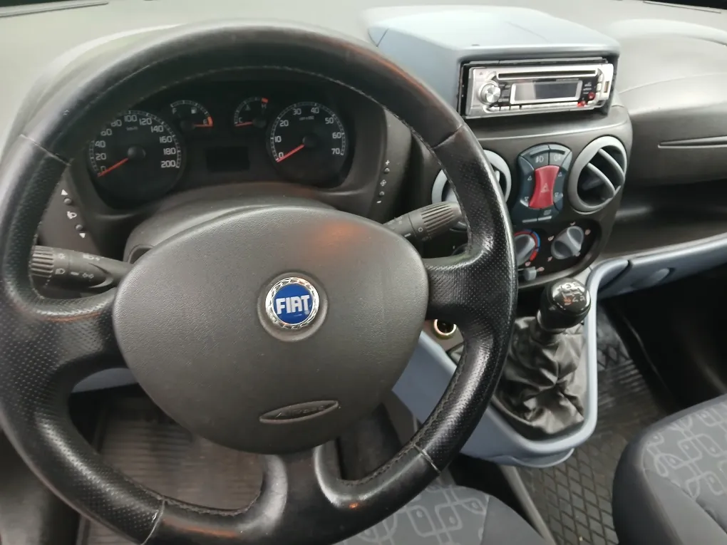 Wnętrze samochodu marki Fiat, widok z perspektywy kierowcy. W centrum znajduje się czarna, skórzana kierownica z logo FIAT na środku. Za kierownicą widoczny jest panel wskaźników z prędkościomierzem i obrotomierzem. Po prawej stronie znajduje się konsola środkowa z radiem, pokrętłami klimatyzacji, nawiewami powietrza oraz lewarkiem zmiany biegów w skórzanej osłonie.