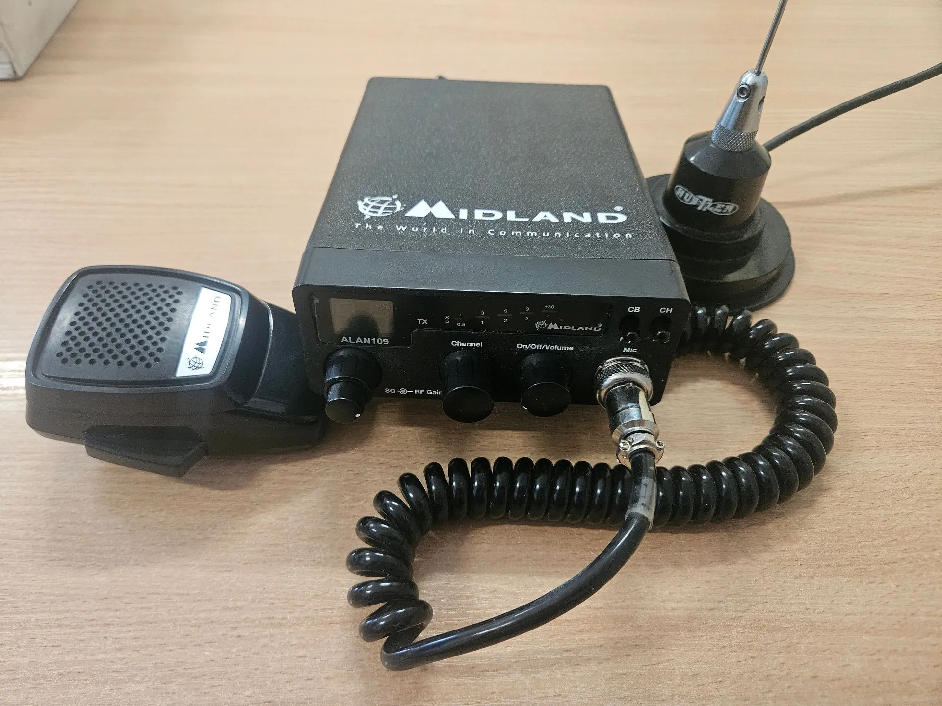 Czarna radiostacja CB marki Midland, model ALAN109, leżąca na jasnym drewnianym blacie. Urządzenie posiada panel przedni z małym wyświetlaczem, kilkoma pokrętłami oraz napisami: 'ALAN109', 'Channel', 'On/Off/Volume', 'SQ RF Gain'. Do radiostacji podłączony jest spiralny kabel prowadzący do mikrofonu ręcznego, który leży po lewej stronie. Po prawej stronie znajduje się podstawa anteny z zamontowanym kablem.