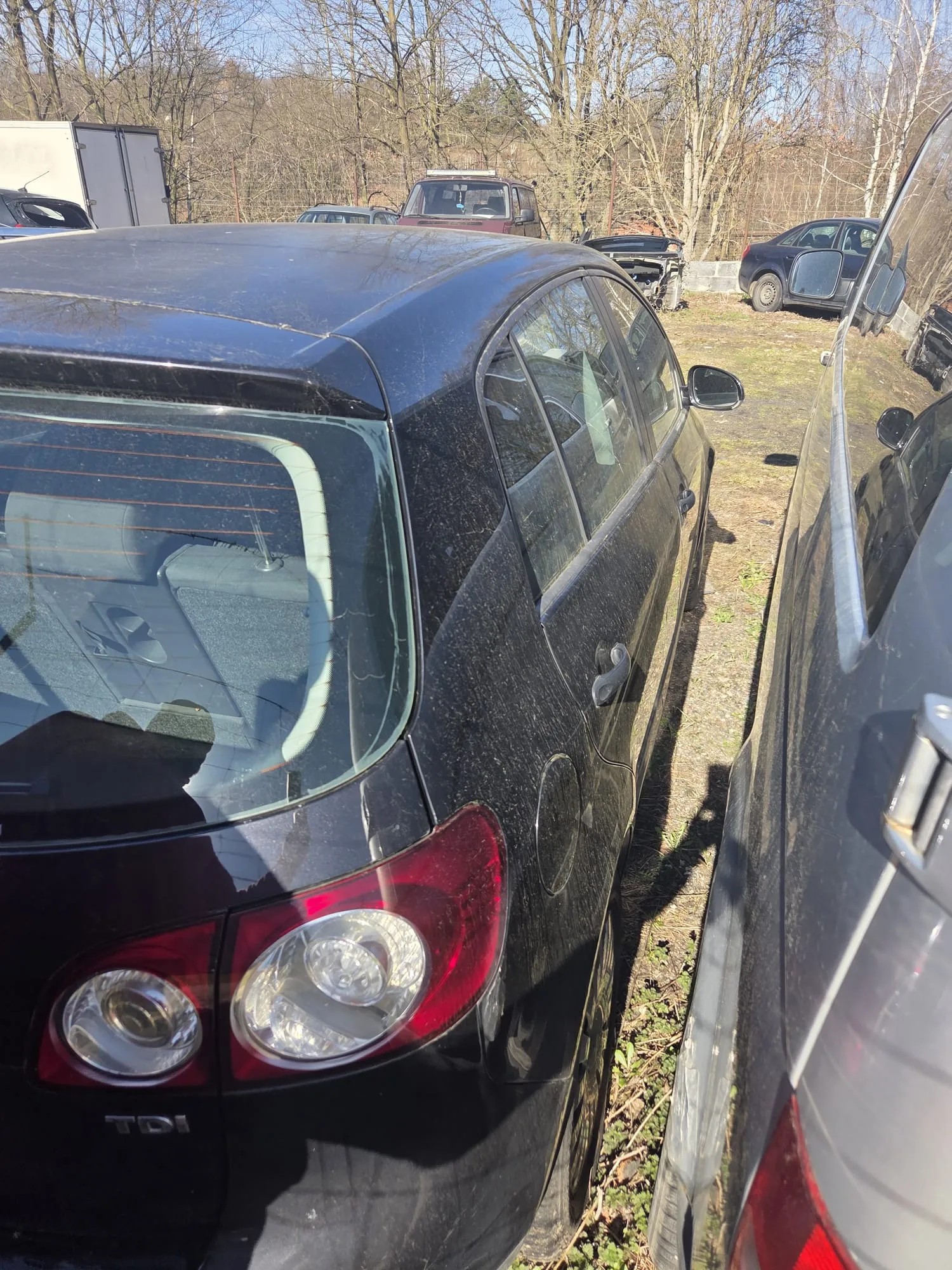 Czarny samochód osobowy typu sedan z widocznym napisem 'TDI' na tylnej części. Widok z tyłu i boku, zaparkowany na terenie z trawą i suchą ziemią. W tle inne pojazdy, drzewa bez liści oraz jasne niebo. Słoneczny dzień.