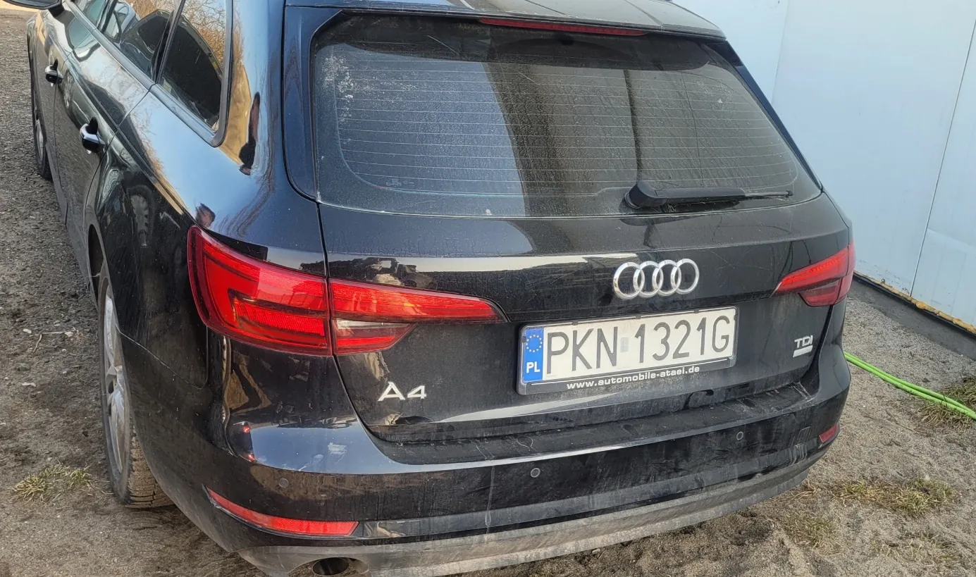 Czarny samochód Audi A4 widziany od tyłu, zaparkowany na podłożu z żwiru. Na tylnej szybie widoczne są ślady brudu. Tablica rejestracyjna z białym tłem i czarnym napisem PKN 1321G, po lewej stronie niebieski pasek z literami PL oraz niebieskim okręgiem z gwiazdkami. Poniżej tablicy napis www.automobile-atael.de. Po lewej stronie bagażnika napis A4, po prawej TDI. W centrum bagażnika logo Audi z czterema połączonymi pierścieniami. Czerwone światła tylnego oświetlenia po obu stronach. W tle biała ściana i zielony wąż na ziemi.