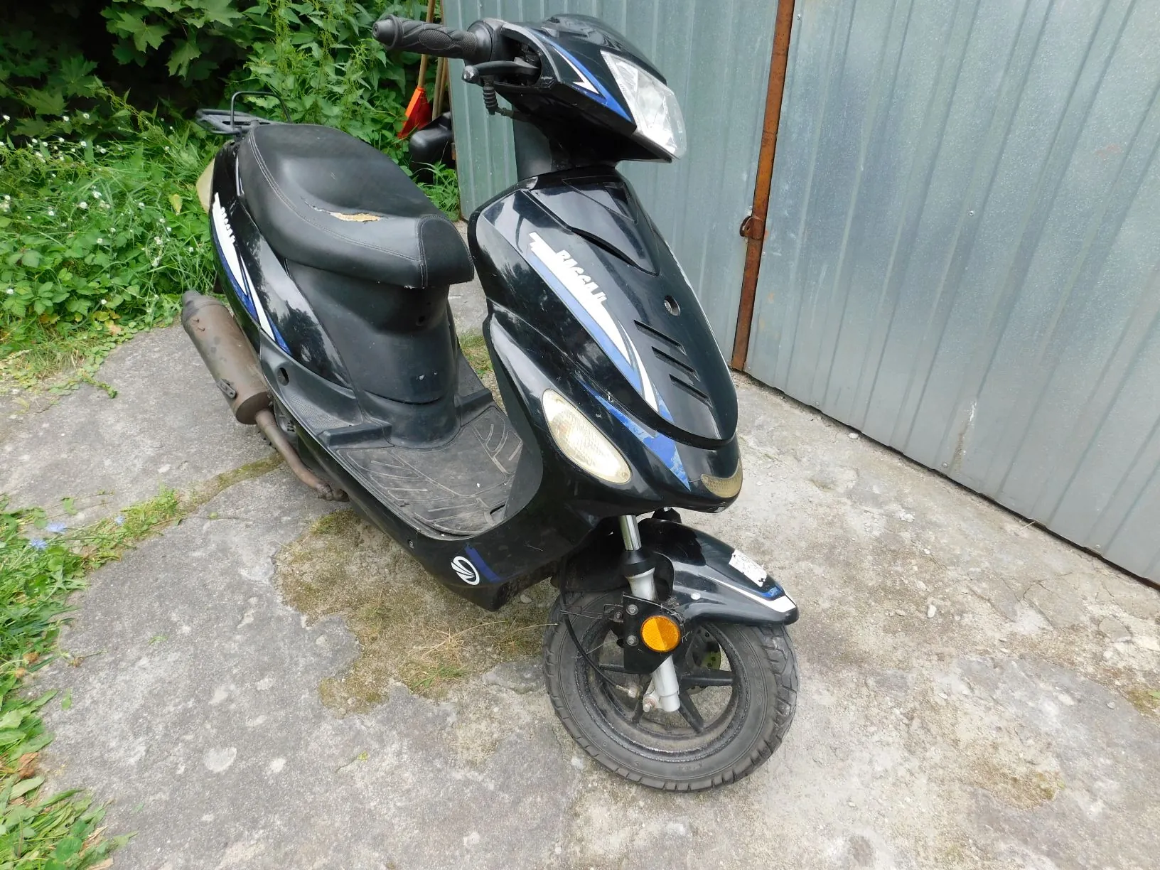 Czarny skuter zaparkowany na betonowym podłożu przed szarym, metalowym ogrodzeniem. Pojazd posiada niebieskie i białe akcenty graficzne oraz napis 'Racer' na przedniej owiewce. W tle widoczna jest zielona roślinność.