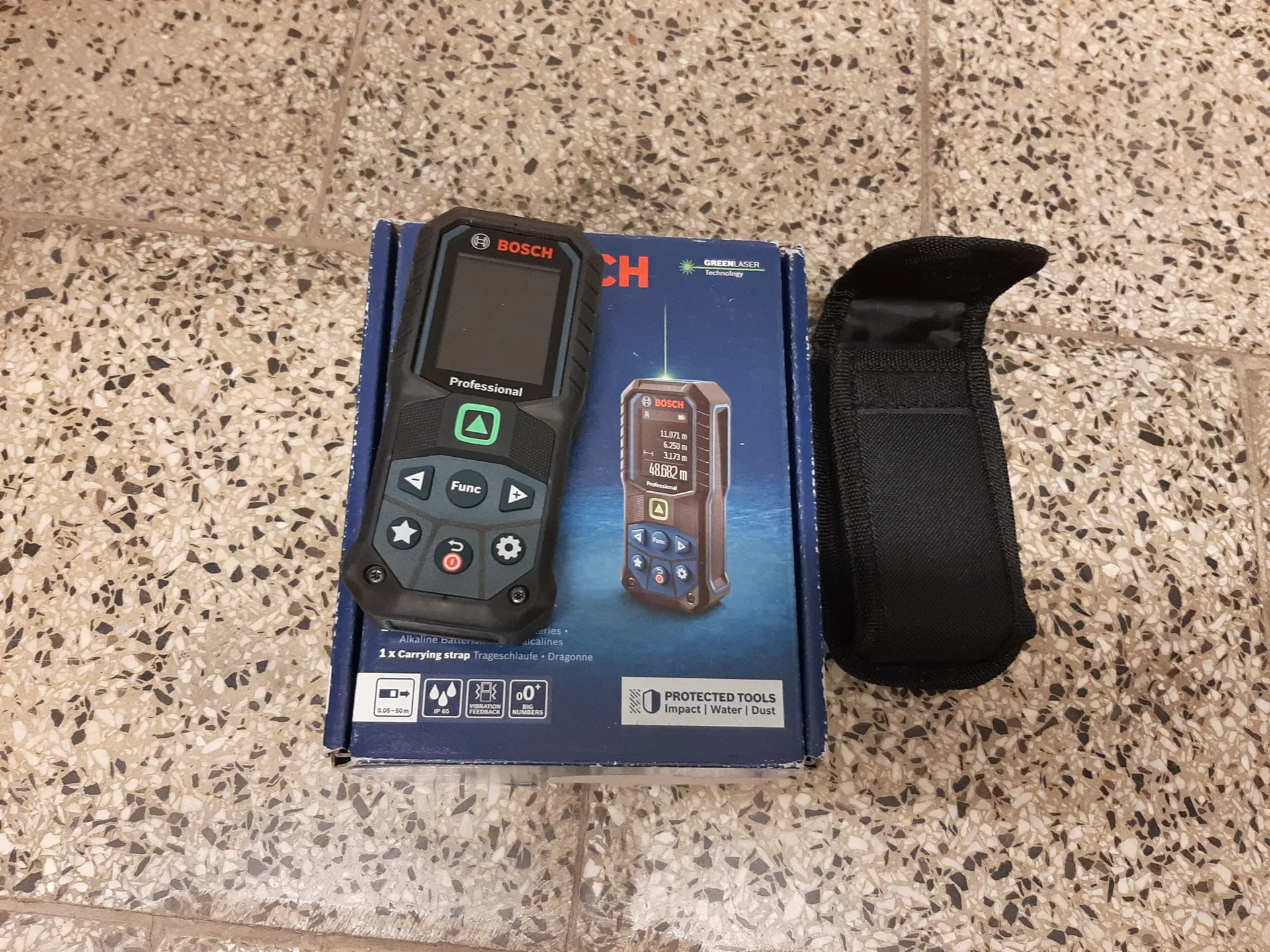 Na zdjęciu widoczny jest czarny dalmierz laserowy marki BOSCH Professional z ekranem i przyciskami (w tym zielonym przyciskiem w kształcie trójkąta, przyciskiem Func oraz innymi symbolami). Urządzenie leży na niebieskim pudełku z logo BOSCH, napisem GreenLaser Technology oraz grafiką przedstawiającą dalmierz z wyświetlonymi danymi pomiarowymi (11,071 m, 6,250 m, 3,173 m, 48,682 m). Na pudełku widoczne są również informacje: 1 x Carrying strap Trageschlaufe • Dragonne oraz ikony wskazujące cechy produktu (zasięg 0,05-50 m, ochrona IP54, wibracyjny feedback, odporność na uderzenia/wodę/pył). Po prawej stronie pudełka znajduje się czarna tkaninowa etui na pasek. Tło stanowi podłoga z kafelkami w kolorze beżowym z czarnymi i białymi kropkami.