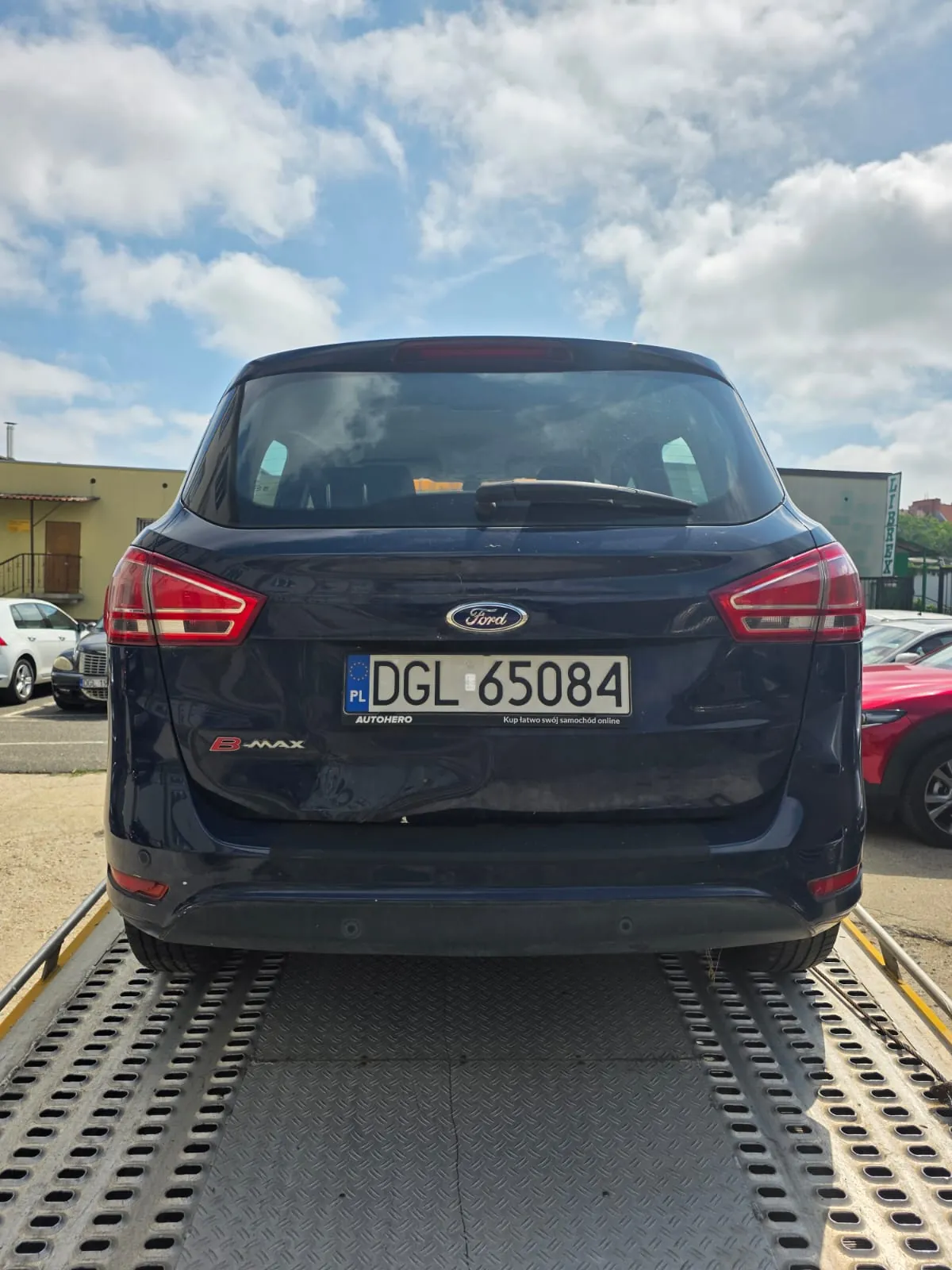 Tylny widok ciemnoniebieskiego samochodu marki Ford B-MAX stojącego na platformie lawety. Samochód posiada polską tablicę rejestracyjną o numerze DGL 65084. Poniżej tablicy widnieje napis AUTOHERO oraz hasło „Kup łatwo swój samochód online”. Po lewej stronie klapy bagażnika znajduje się oznaczenie modelu B-MAX. W tle widoczne jest jasne niebo z chmurami oraz fragmenty innych zaparkowanych pojazdów.