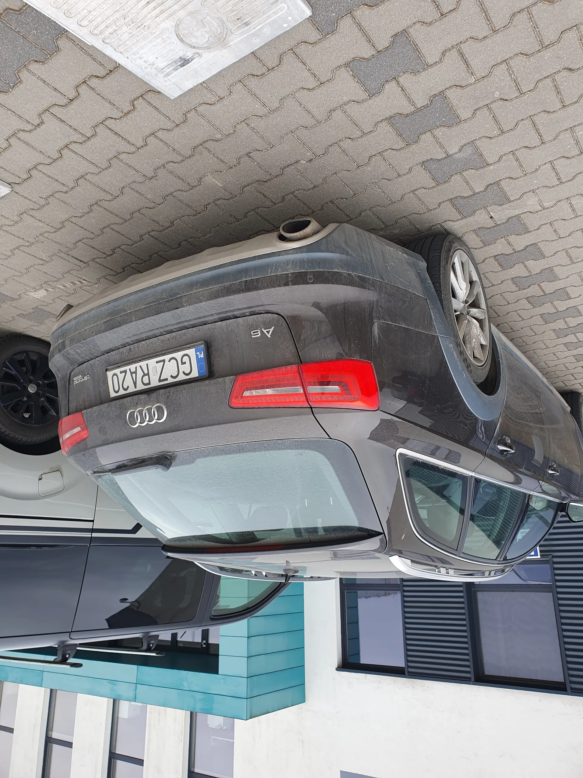 Tylne widok szarego samochodu Audi A6 z tablicą rejestracyjną GCZ RA20 zaparkowanego na brukowanej powierzchni. W tle widoczny fragment białego budynku z turkusowymi elementami oraz częściowo widoczny biały samochód.