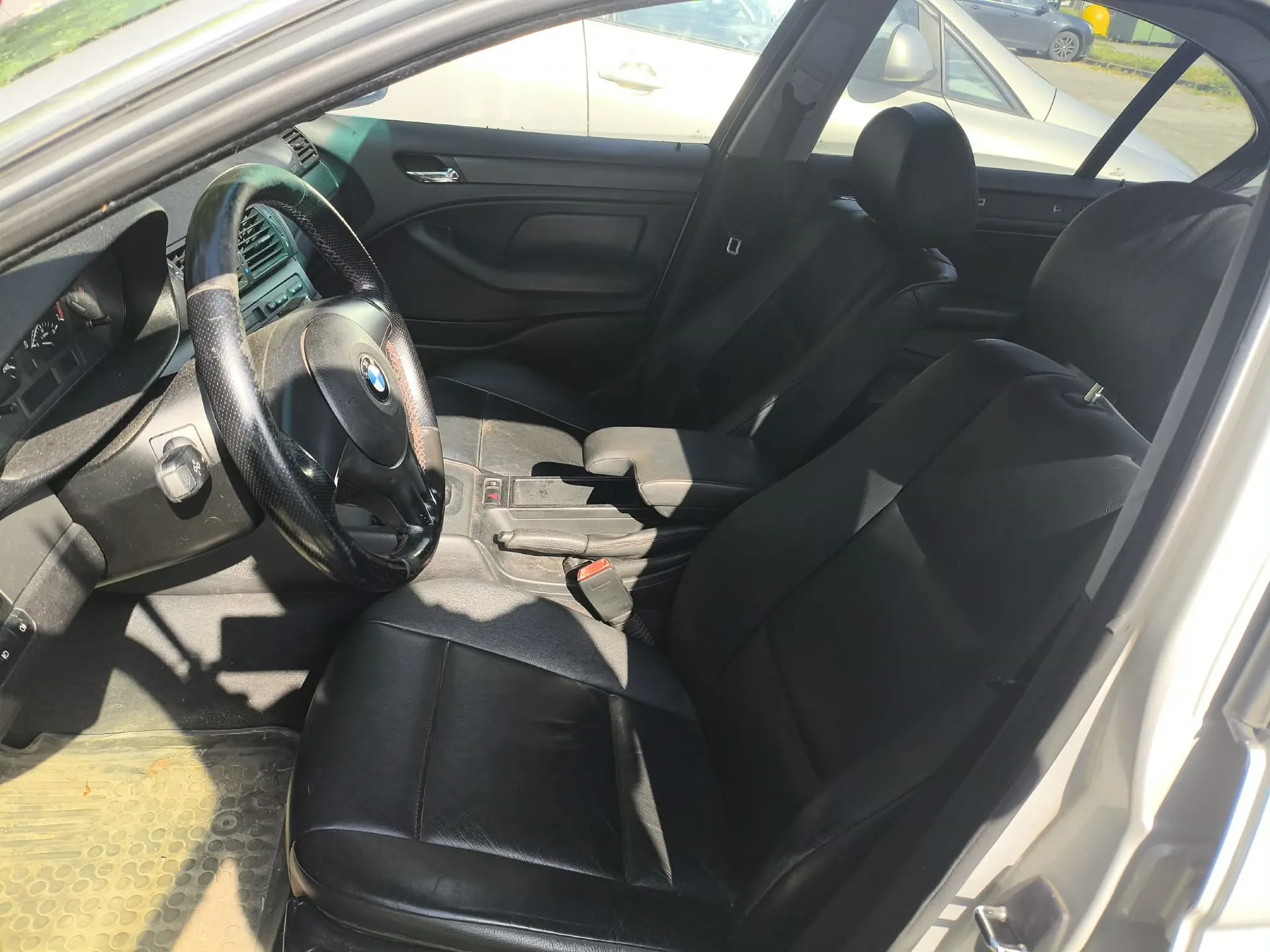 Wnętrze samochodu widziane z perspektywy otwartych drzwi pasażera. Widoczna jest czarna skórzana tapicerka foteli, kierownica z logo BMW, deska rozdzielcza oraz konsola środkowa. Przez okna widać fragmenty innych białych samochodów zaparkowanych na zewnątrz.