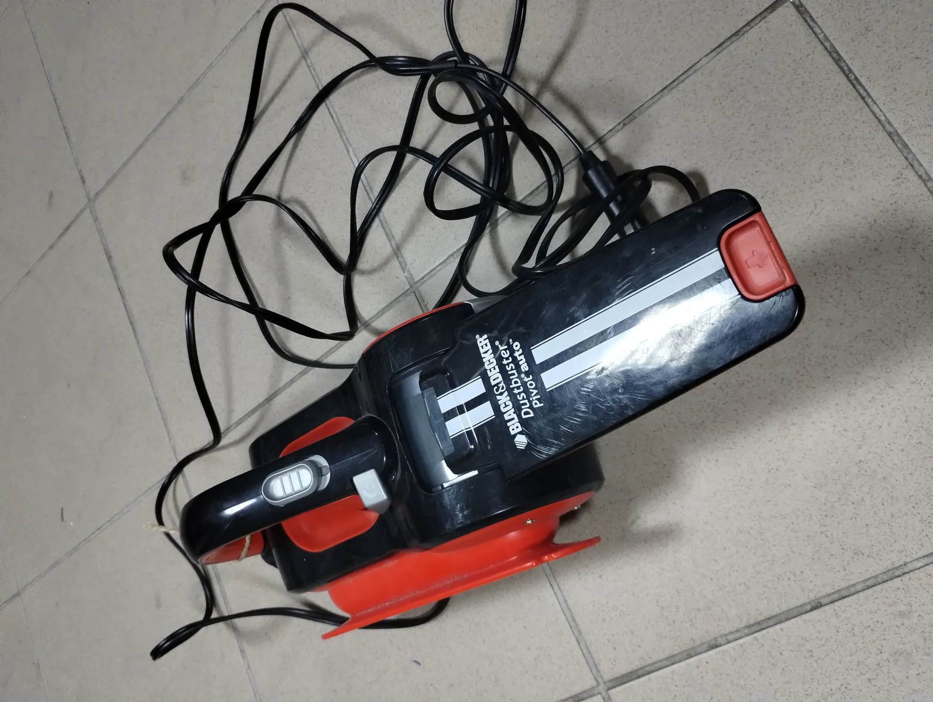 Czarno-czerwony odkurzacz ręczny marki Black & Decker, model Dustbuster Pivot, leżący na jasnej, płytkowej podłodze. Urządzenie ma czarną obudowę z czerwonymi elementami i białym pasem z napisem 'BLACK & DECKER Dustbuster Pivot'. Do odkurzacza przyłączony jest długi, czarny kabel zasilający, który jest splątany nad urządzeniem.