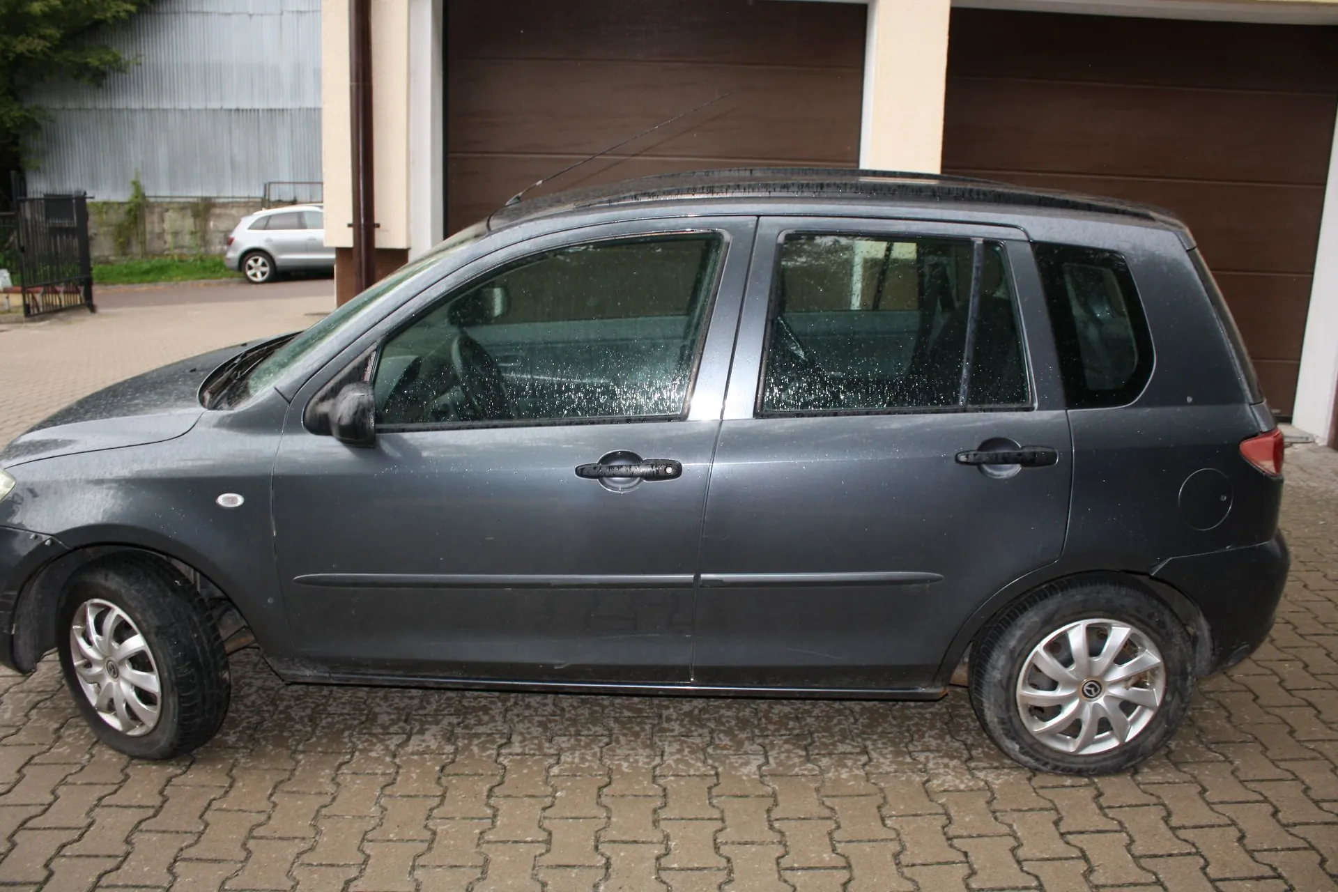 Szary samochód osobowy typu hatchback zaparkowany bokiem na kostce brukowej przed brązową bramą garażową. Na karoserii auta widoczne są krople wody. W tle, po lewej stronie, widać fragment innego, srebrnego samochodu oraz metalowy płot.