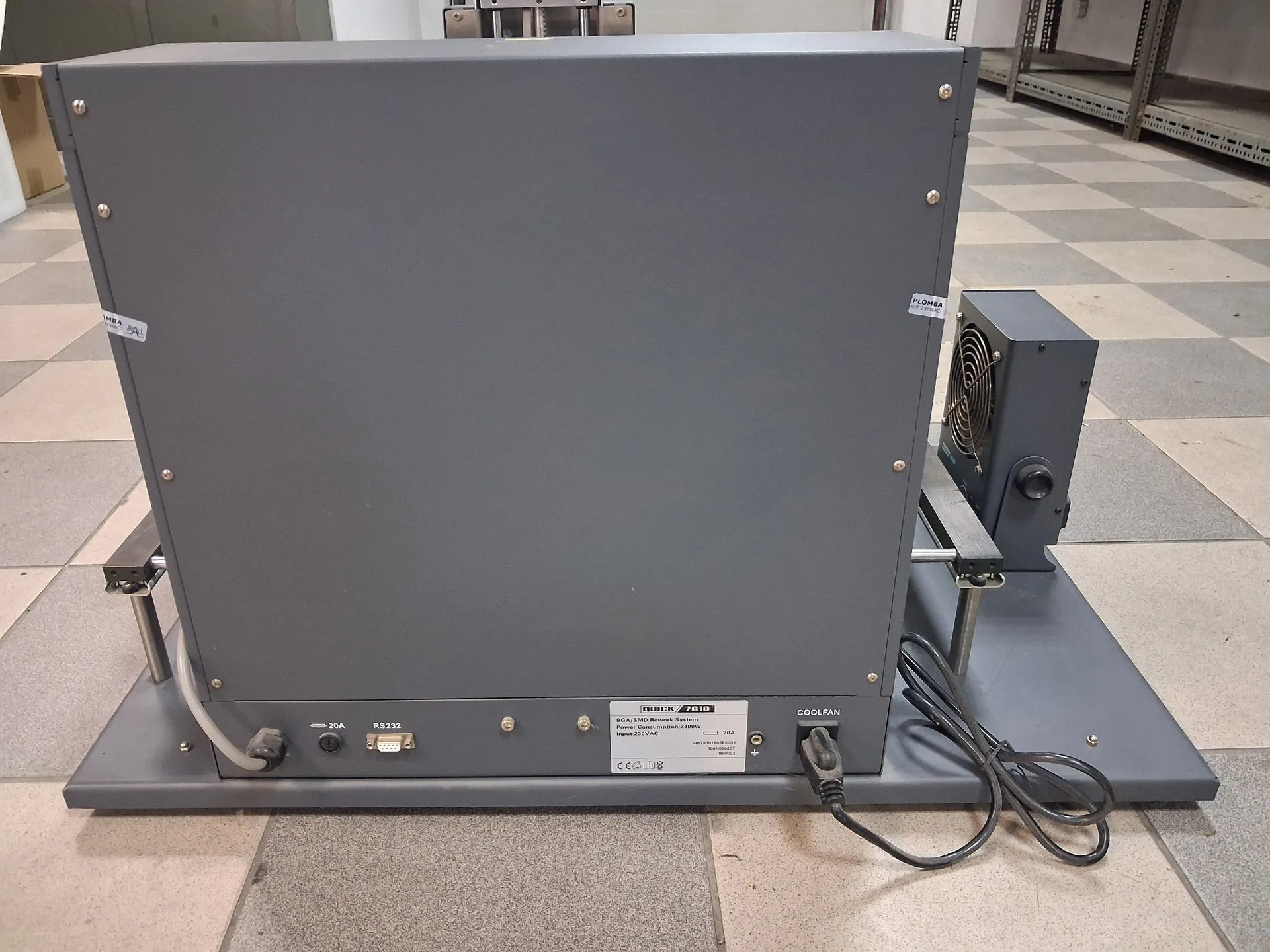 Szary, metalowy moduł elektroniczny w obudowie w kształcie prostopadłościanu, ustawiony na płaskiej podstawie. Na tylnym panelu urządzenia znajduje się tabliczka znamionowa z tekstem: 'BIRCH 2610', 'BCA/SMB Automatic System', 'Power: 3-phase 380VAC 2400W', 'Input: 230VAC'. Widoczne są również gniazda RS232, przełącznik zasilania 20A oraz podłączony kabel zasilający. Po prawej stronie obudowy zamontowany jest zewnętrzny wentylator chłodzący (COOLFAN). Urządzenie stoi na jasnej, kafelkowej podłodze.