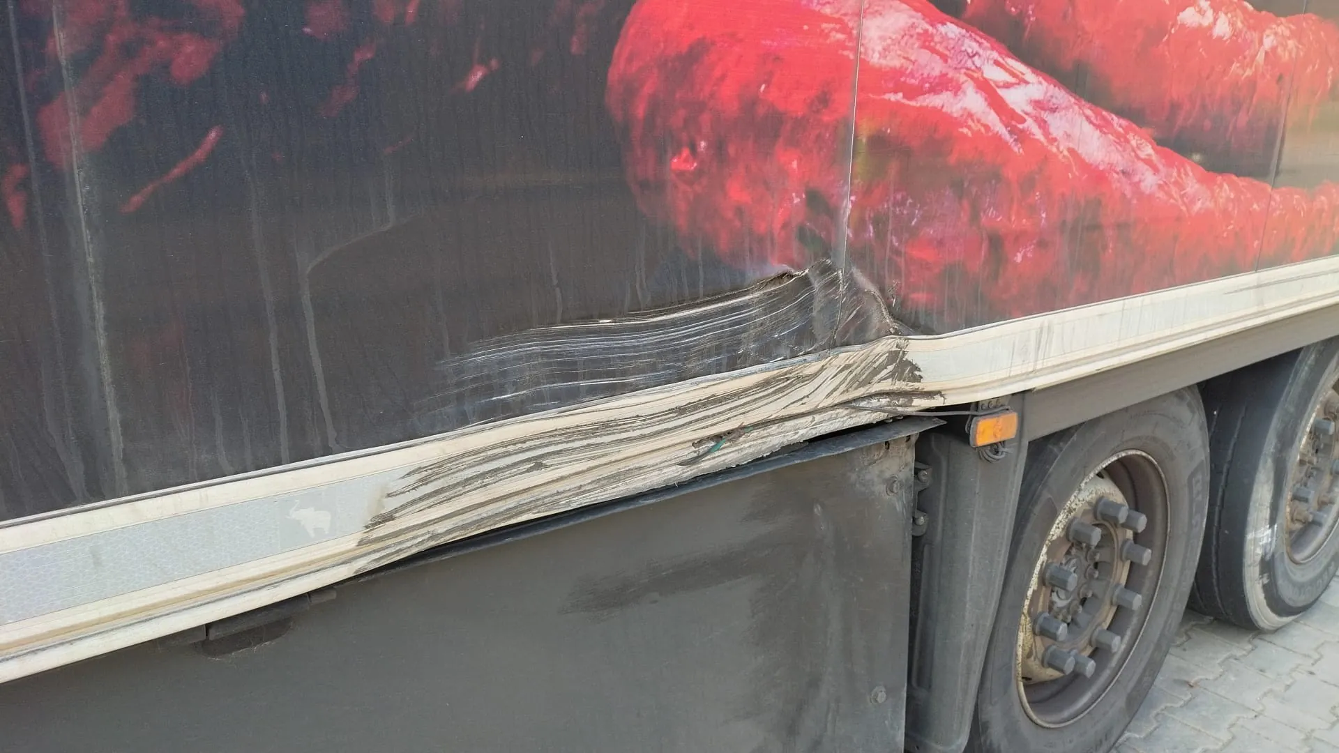 Fragment boku ciężarówki z uszkodzonym nadwoziem. Widoczne są duże czerwone papryki na grafice reklamowej, zarysowane ślady uszkodzenia metalowego pasa bocznego oraz koła z widocznymi śrubami i częścią podwozia. Tło stanowi brukowana powierzchnia.
