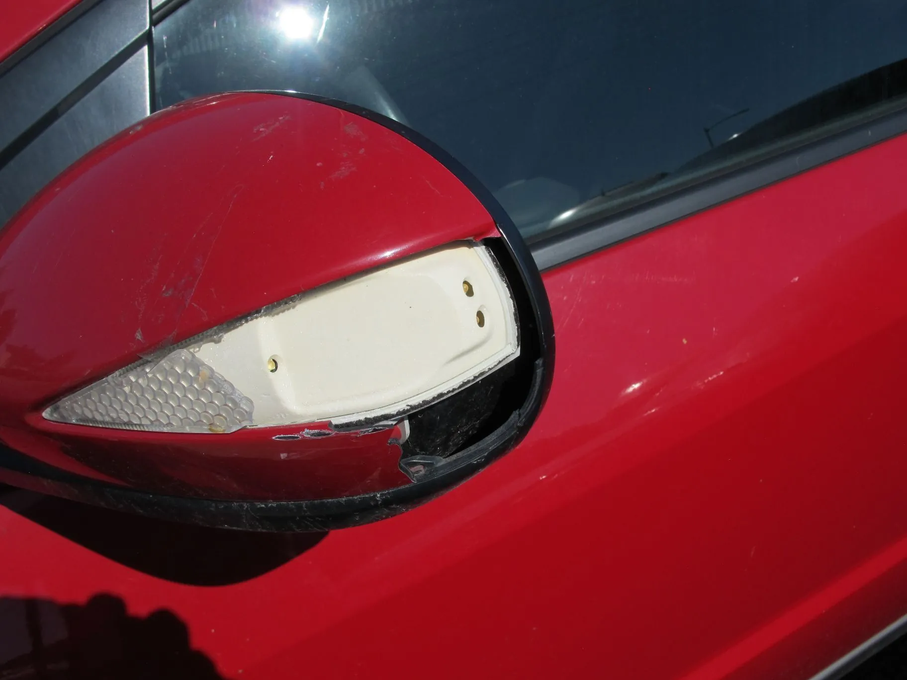 Zbliżenie na uszkodzoną, czerwoną obudowę lusterka bocznego w samochodzie. Plastikowa czerwona osłona jest pęknięta i częściowo oderwana, odsłaniając biały element wewnętrzny oraz przezroczystą soczewkę kierunkowskazu o strukturze plastra miodu.